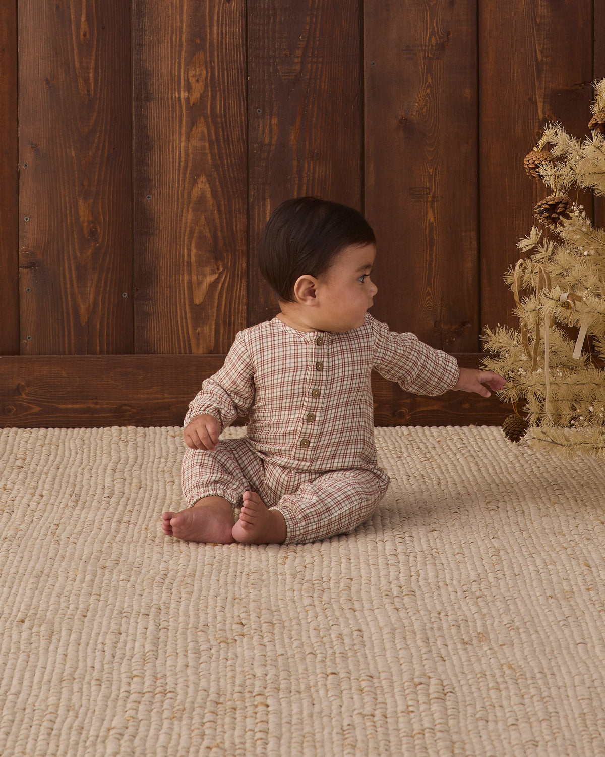 A baby sitting on a textured cream rug in front of a wood-paneled wall, wearing a long-sleeve Christmas plaid jumpsuit with a button placket on the chest.