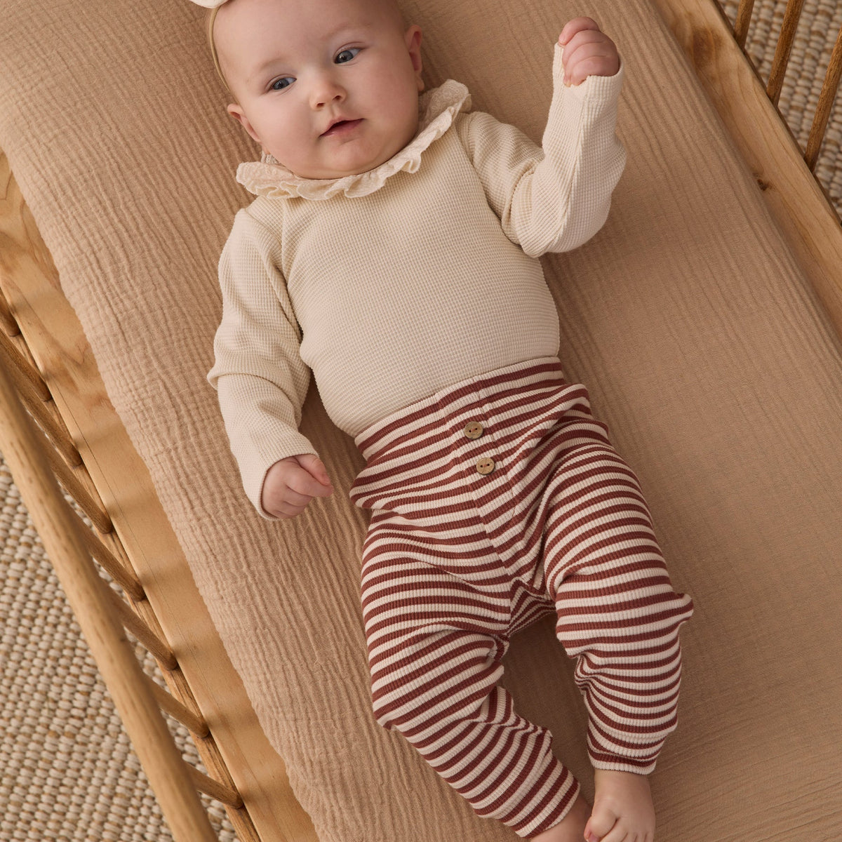 A baby lying on a woven bassinet, wearing a long-sleeve natural beige ruffled collar bodysuit and brown-and-white striped pants, wearing a small hair bow.