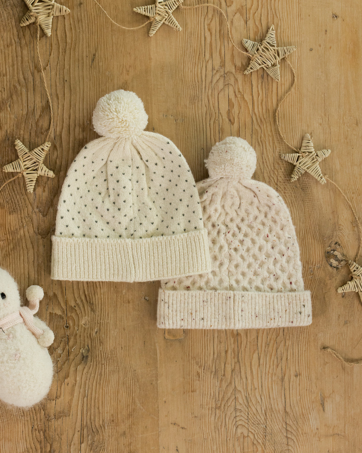 Two cream-colored knit baby hats with pom-poms on a wooden surface with decorative stars.