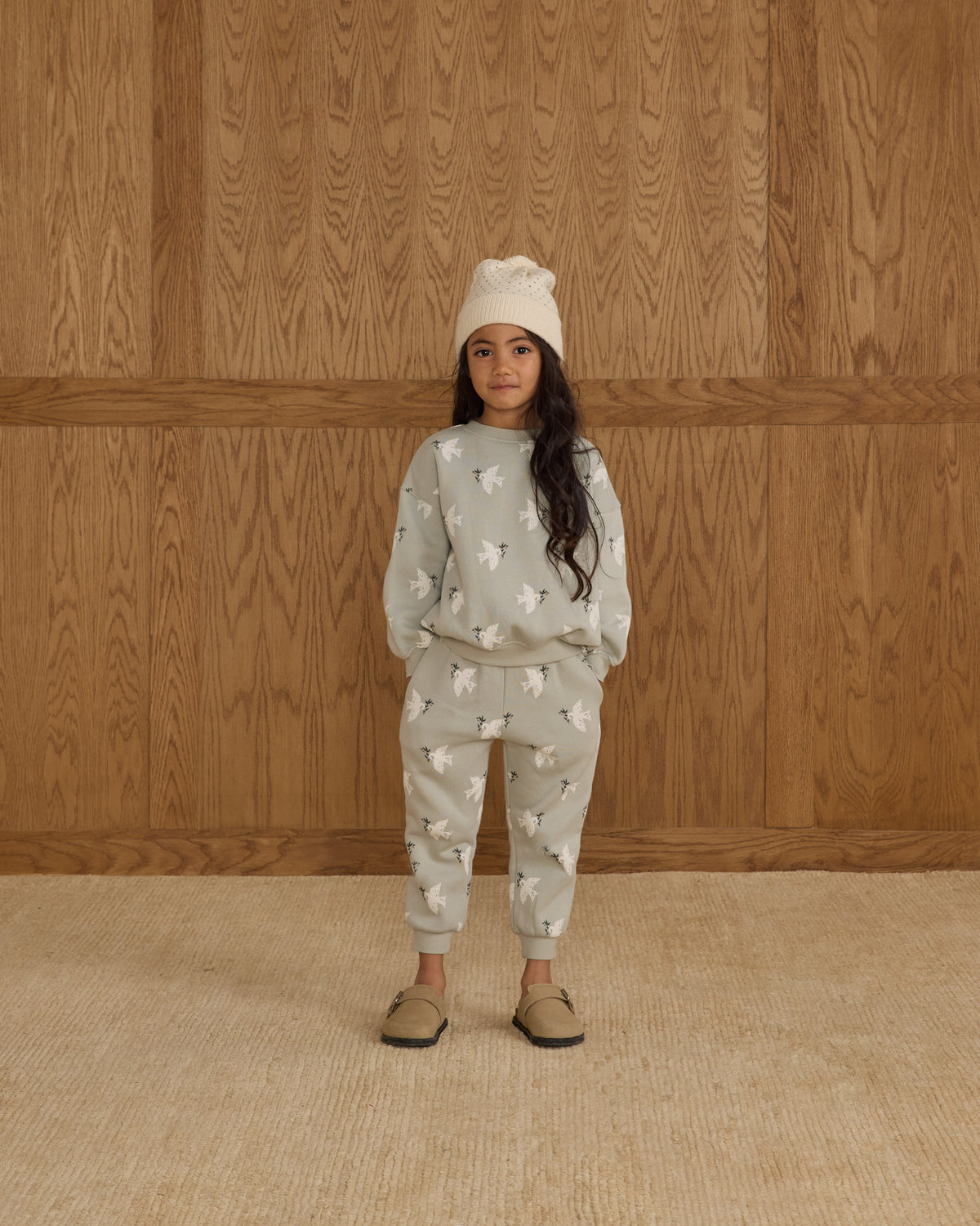 Child wearing a light gray pajama set with a pattern against a wooden paneled wall.
