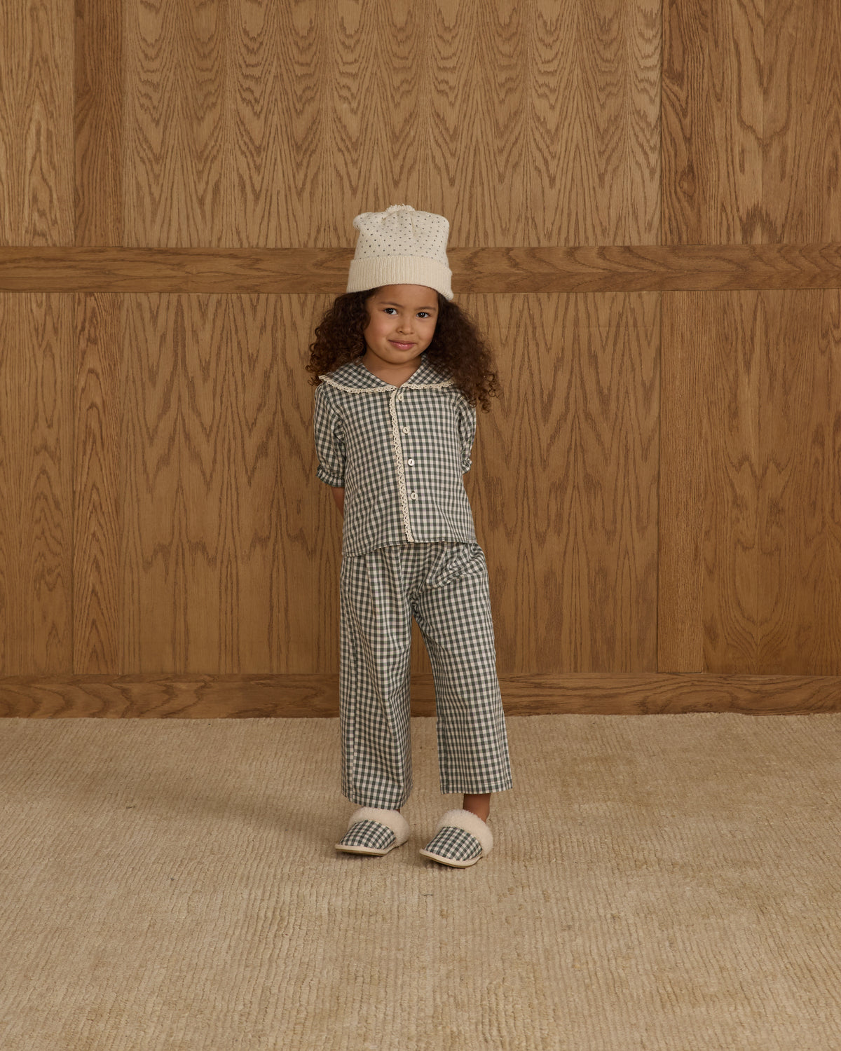 Child wearing a checkered outfit with a white hat against a wooden paneled wall.