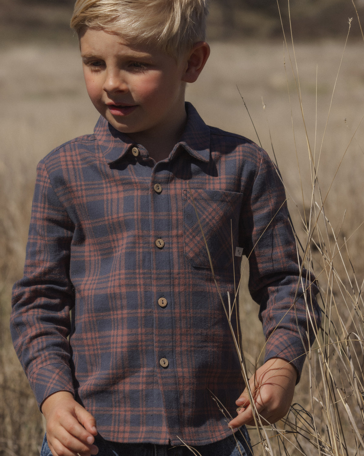 A young boy wearing a long-sleeve button-up plaid shirt in navy and red, standing in a dry grassy field.