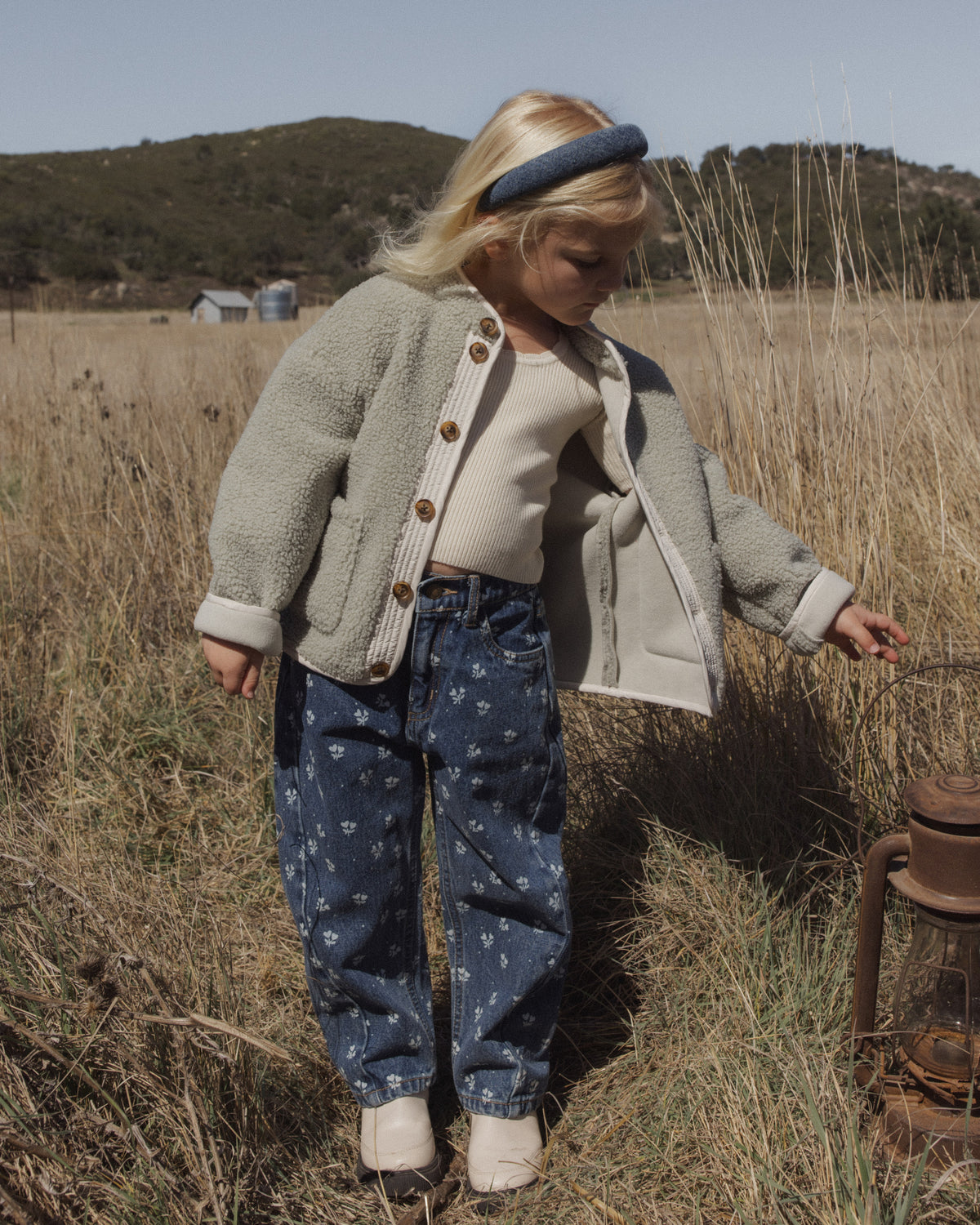 A young child standing in a dry grassy field outdoors, wearing a light beige sherpa button-front cardigan over a cream top, blue patterned pants, and a dark blue headband.