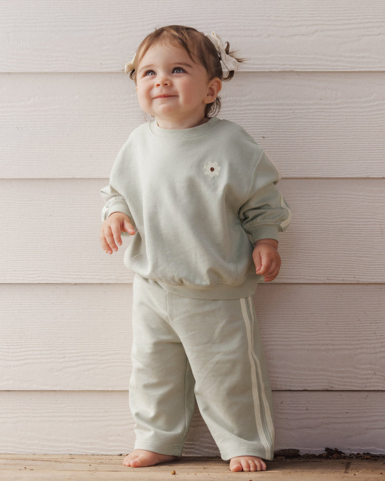 A toddler wearing a sage-green relaxed sweatshirt and matching sweatpants set with side-stripe detailing from Rylee + Cru.
