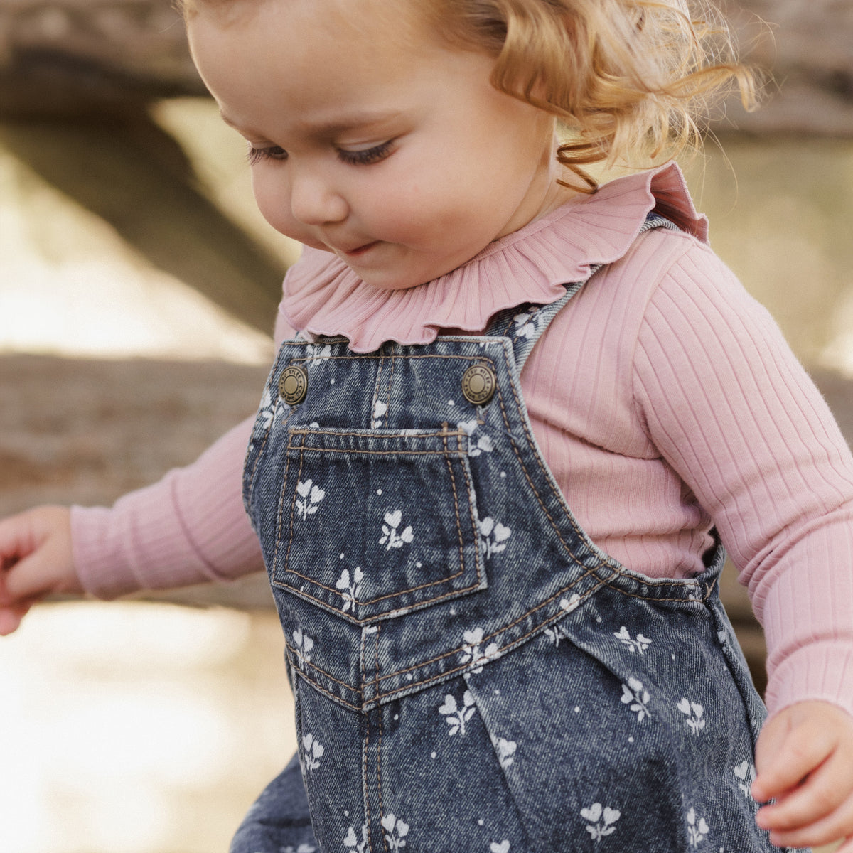 A toddler girl wearing a dusty pink ribbed long-sleeve top and dark blue denim overall with a white floral pattern, looking down in an outdoor setting.