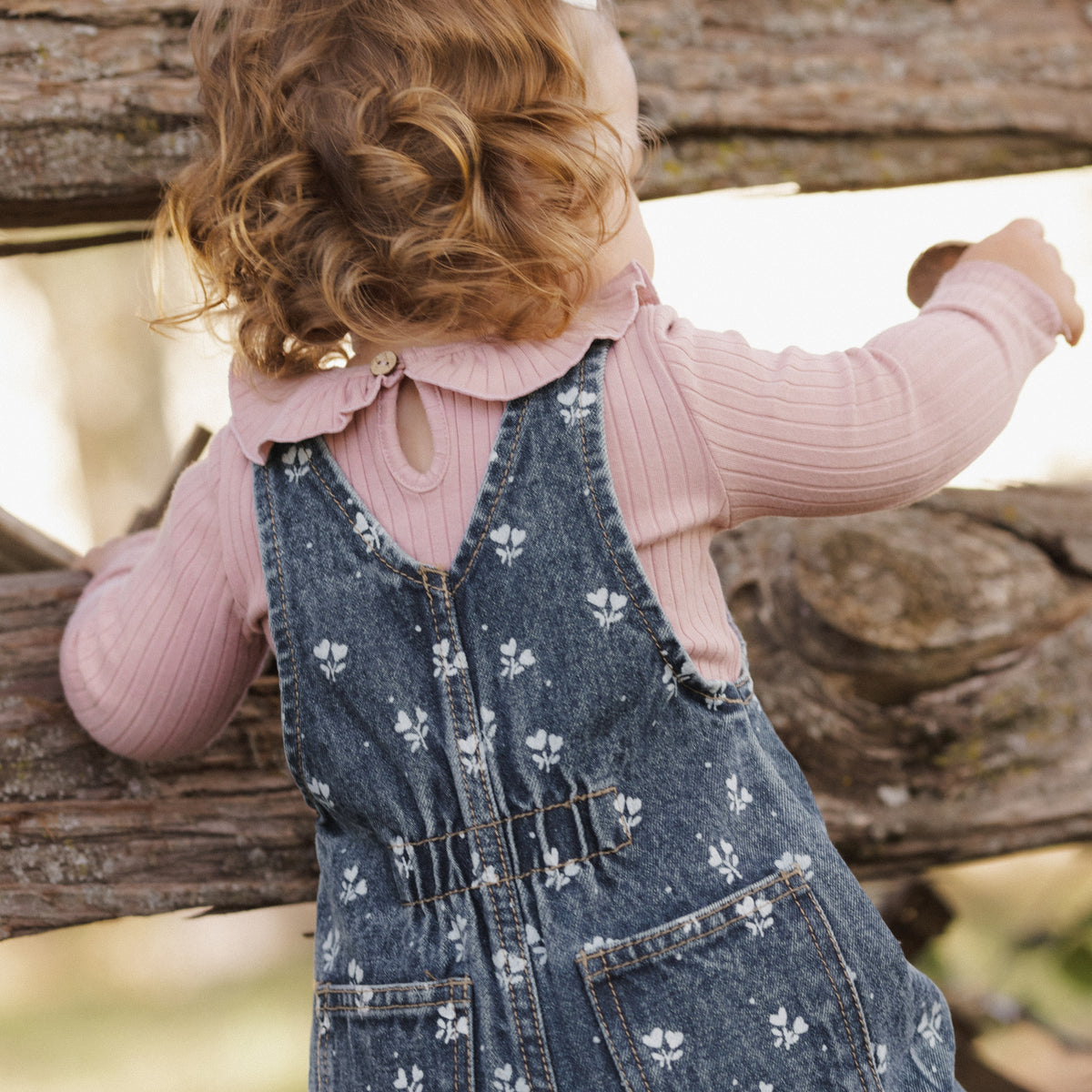 A toddler seen from behind wearing a pink ribbed long-sleeve top and a blue denim overall with a white floral print, leaning on a rustic wooden fence.