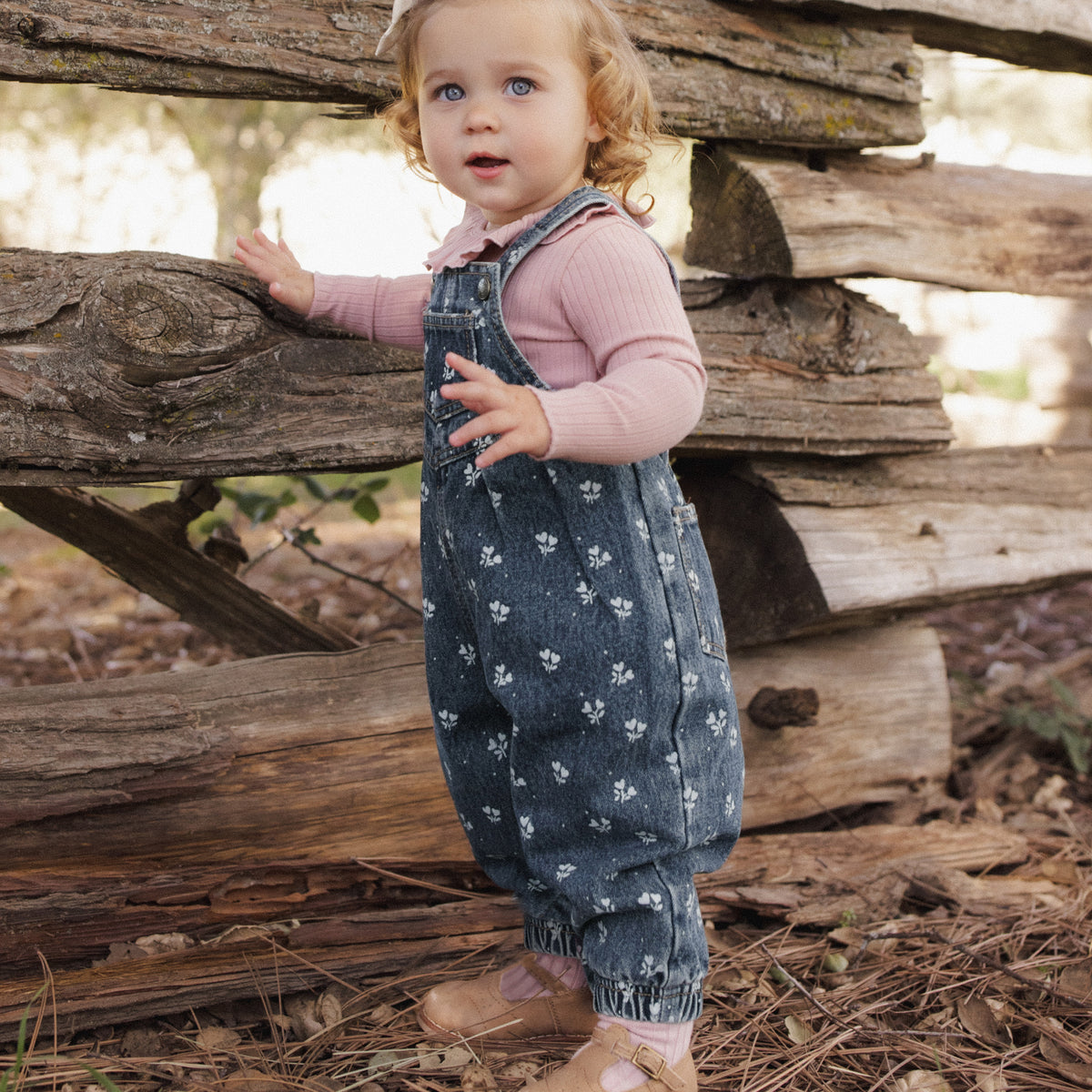 A toddler girl wearing a pink long-sleeve top and dark blue patterned overall-style romper stands outdoors in front of a rustic wooden fence on a wooded path.