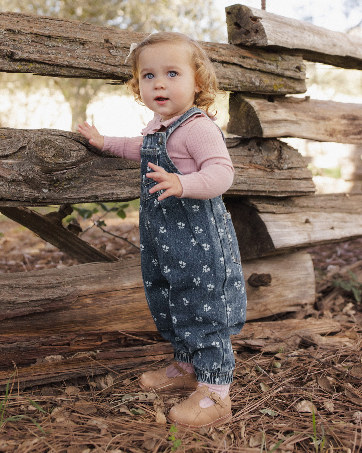 A toddler girl wearing a pink long-sleeve top and dark blue patterned overall-style romper stands outdoors in front of a rustic wooden fence on a wooded path.