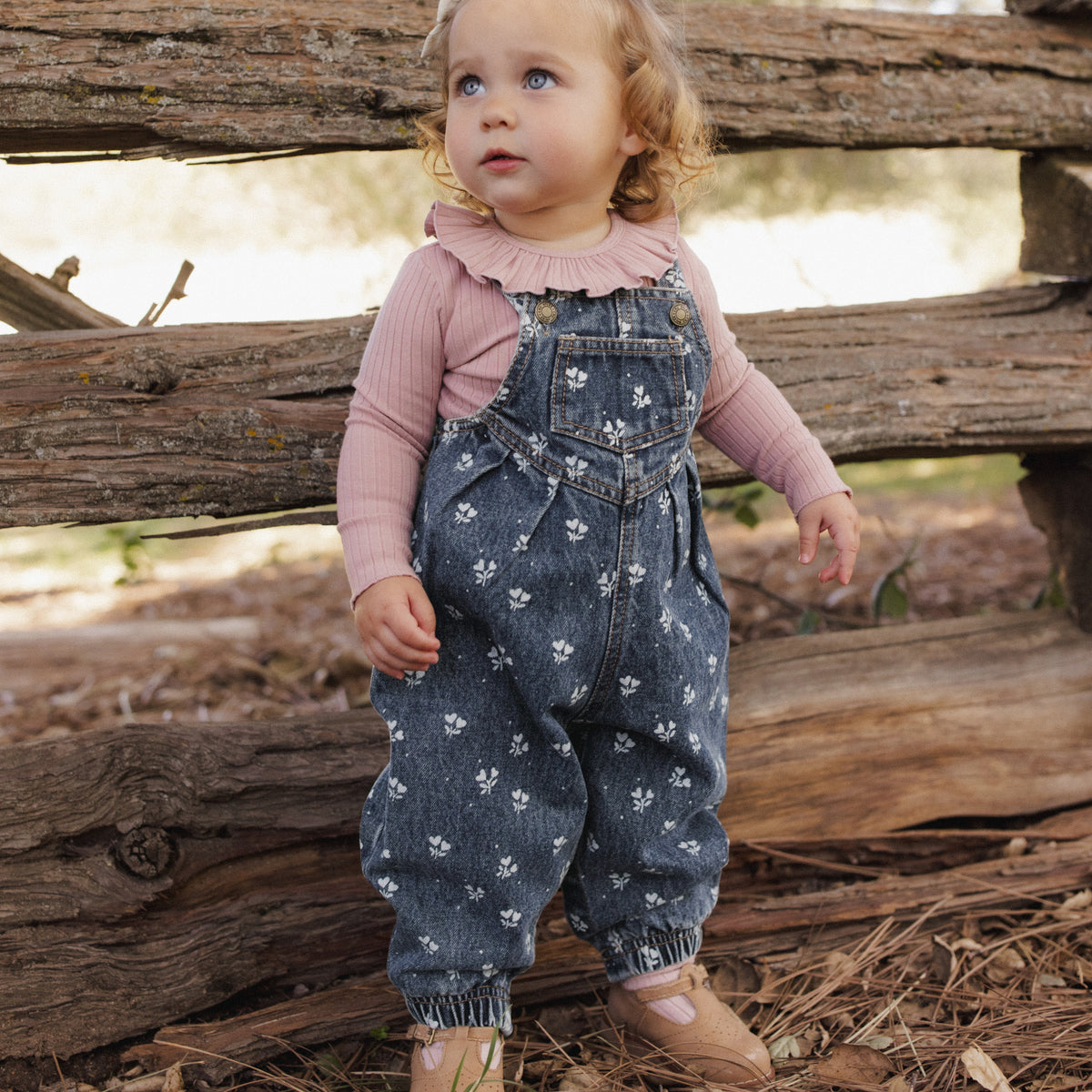A toddler standing outdoors in front of a rustic wooden fence, wearing blue denim overall with a white floral print over a dusty pink long-sleeve ruffled shirt and tan shoes.