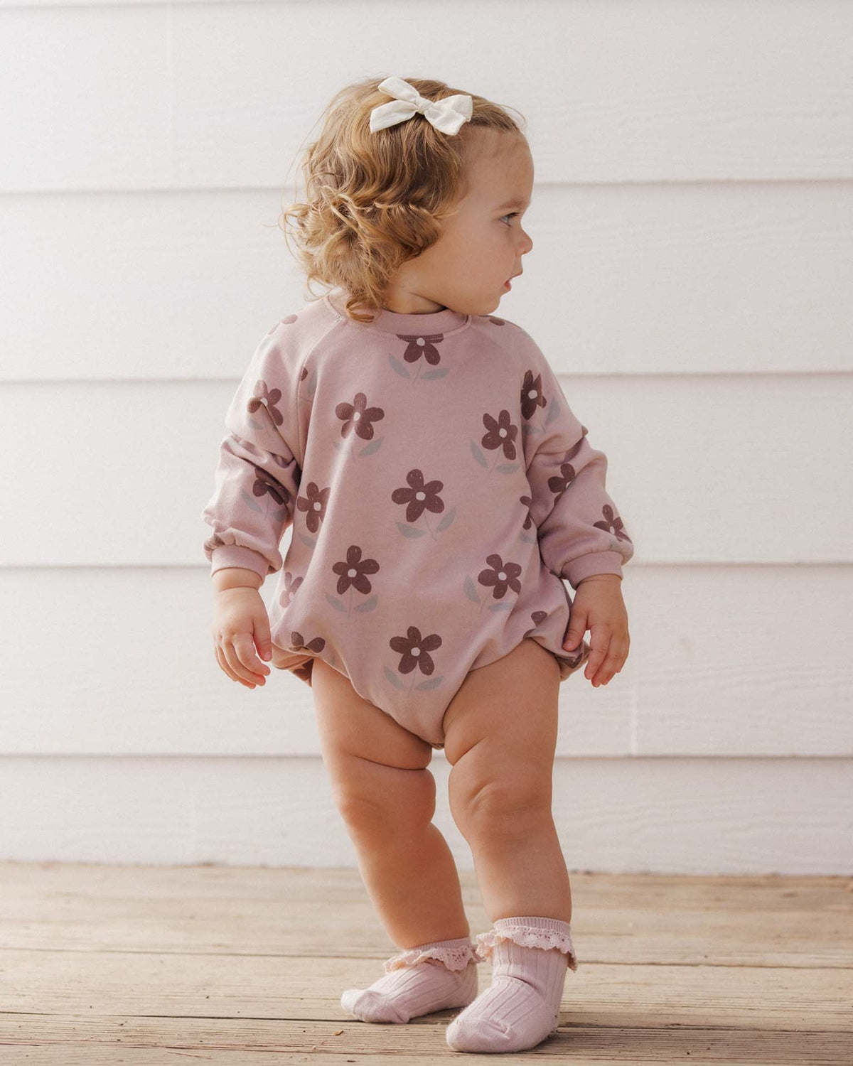 A toddler wearing a mauve-pink long-sleeve crewneck bubble romper featuring an all-over brown daisy floral print by Rylee + Cru.