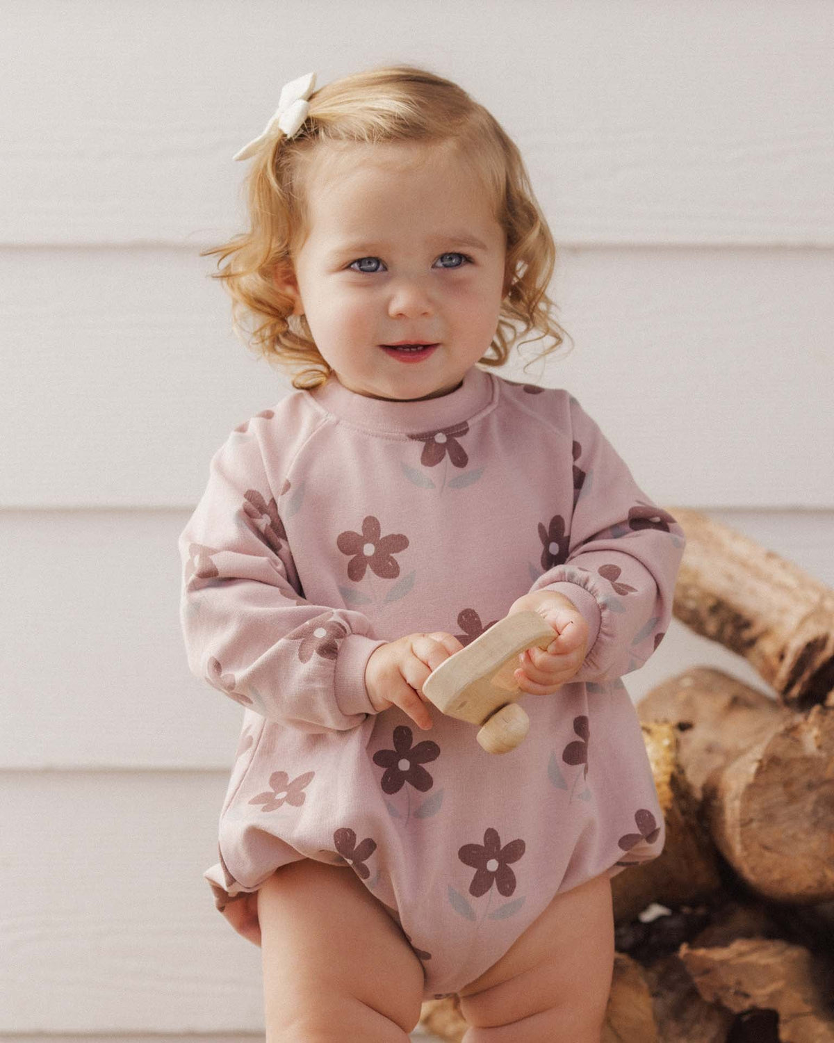 A toddler wearing the Rylee + Cru Daisy Crew Neck Romper in a soft pink hue with an all-over dark floral print.