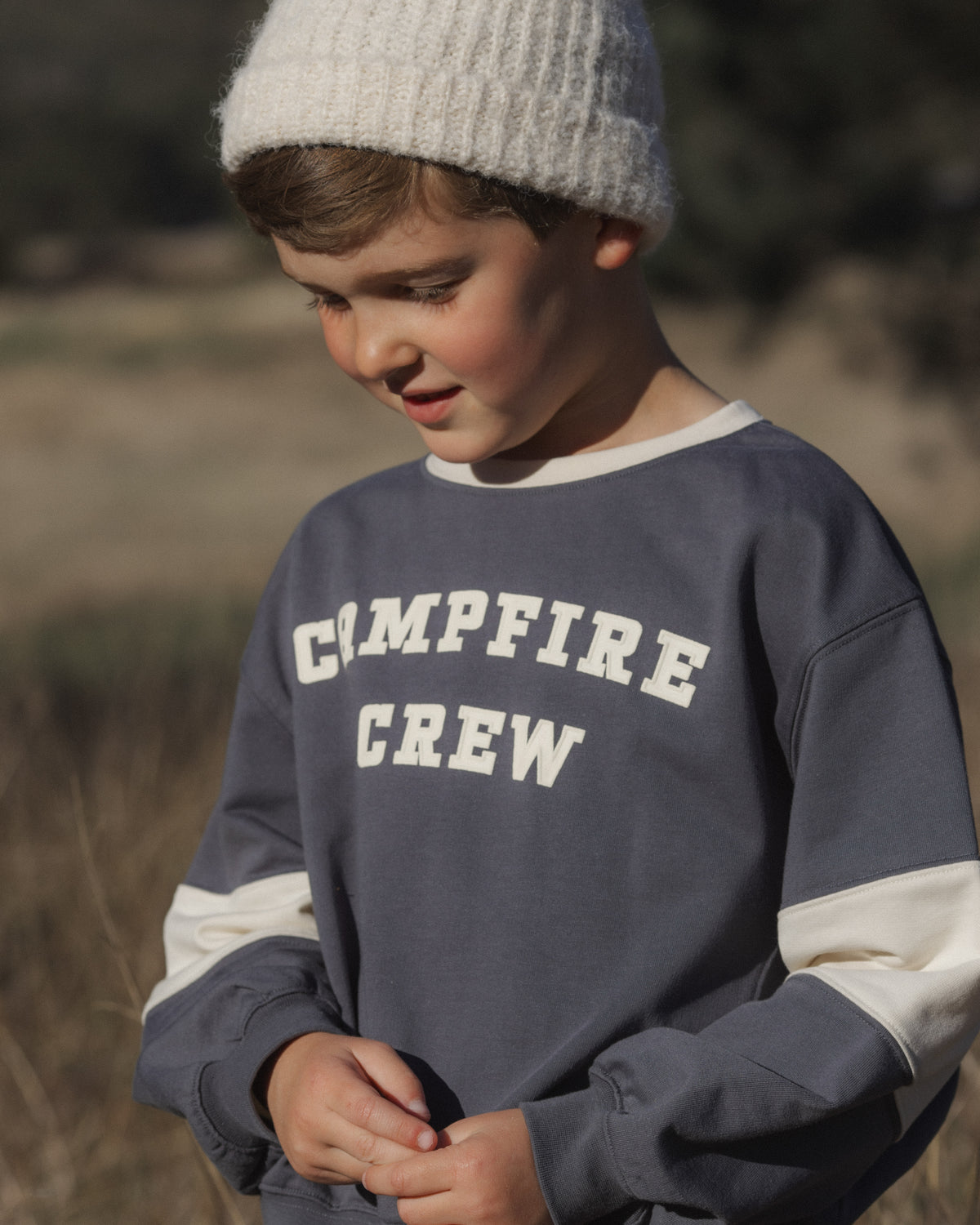 A young boy wearing a slate blue crewneck sweatshirt with cream-colored sleeve panels and 'CMPFIRE CREW' printed in cream on the chest, paired with a cream knit beanie, set against a blurred outdoor background.