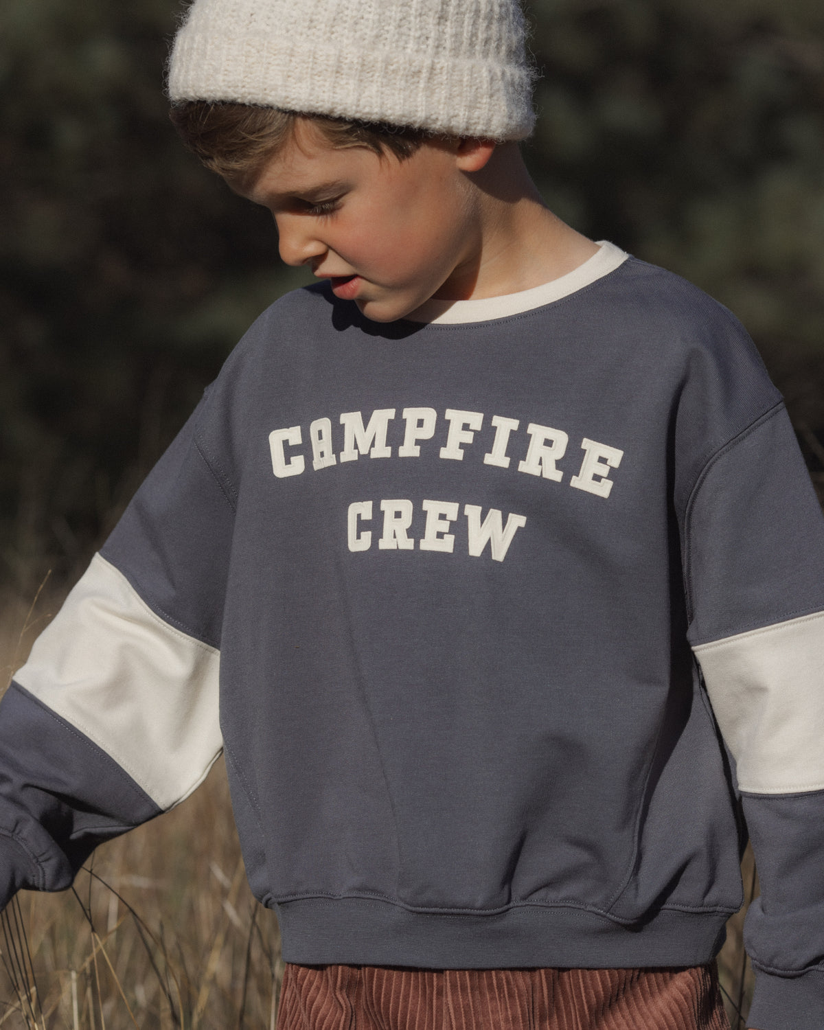 A child wearing a slate blue and white color-block sweatshirt with 'CAMPFIRE CREW' printed in white varsity lettering on the chest, paired with a white knit beanie.