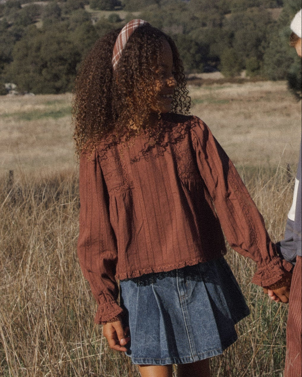 A person standing outdoors in a dry grassy field wearing a rust-brown long-sleeve textured blouse with lace trim at the neckline, paired with a blue denim skirt.
