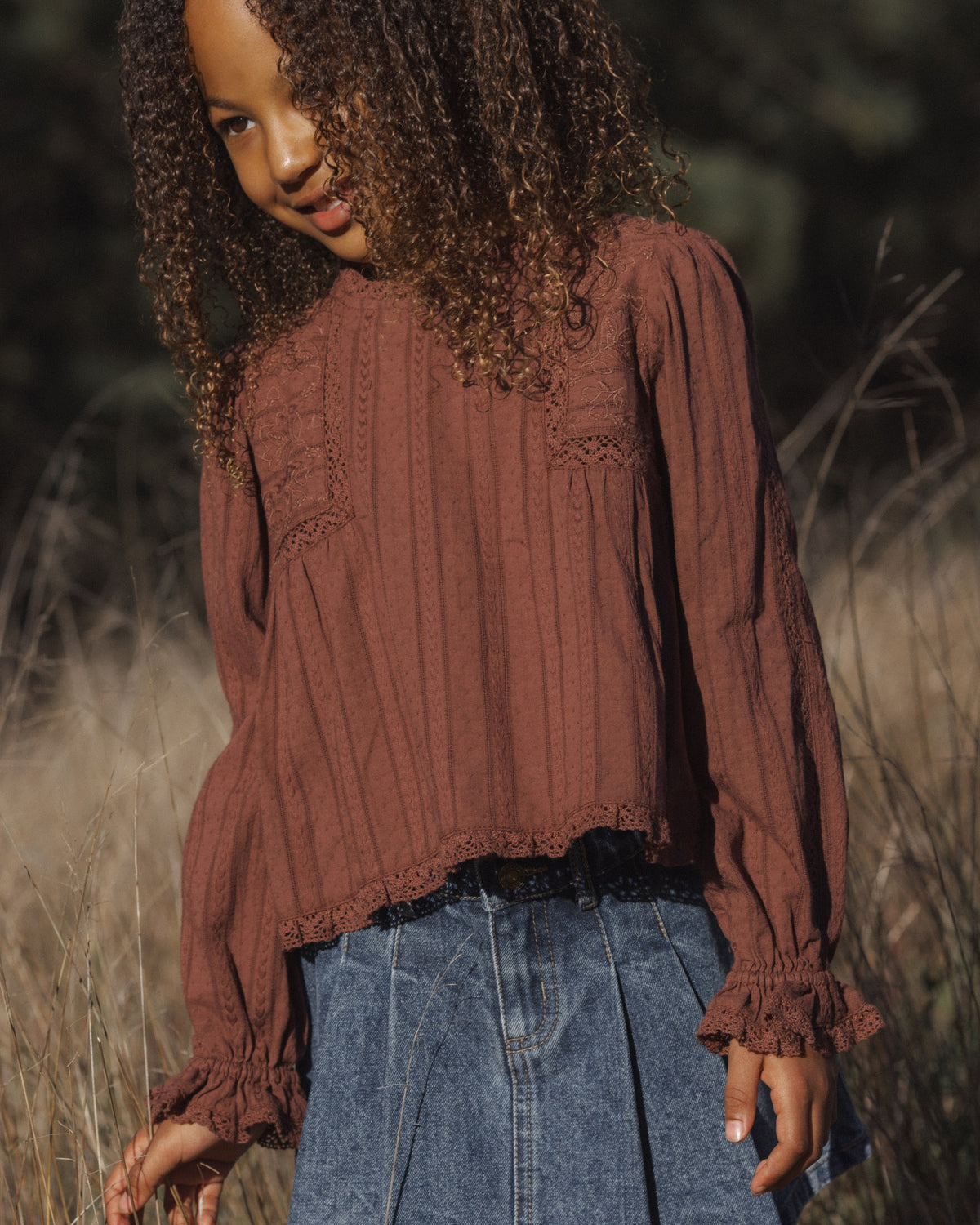 A woman wearing a rust-brown long-sleeve pleated blouse and blue jeans standing outdoors in a field of dry grass.