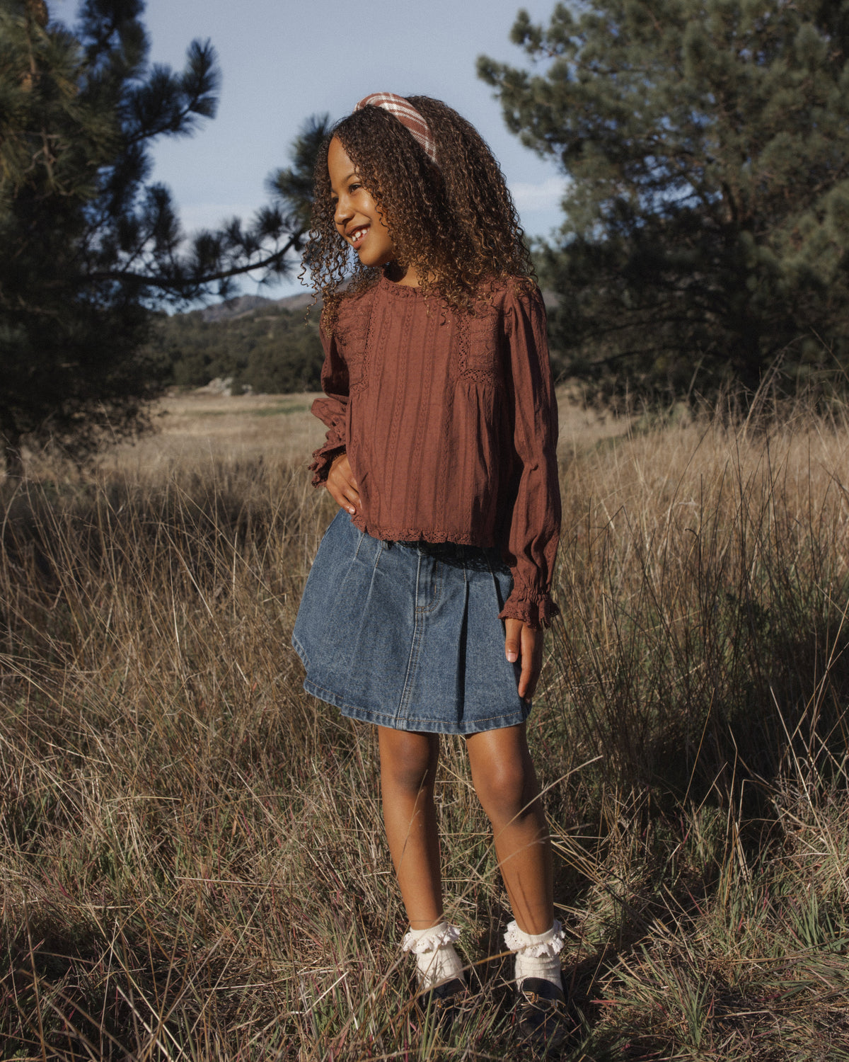 A young girl standing outdoors in a grassy field, wearing a rust-brown long-sleeve top, a blue denim skirt, and white sneakers.