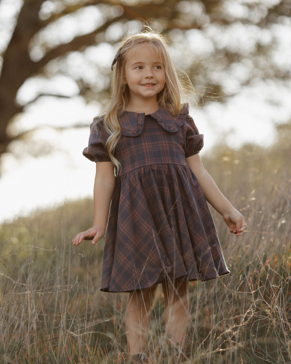 A young girl with long blonde hair stands in a field wearing a dark brown and red plaid dress with a ruffled Peter Pan collar.