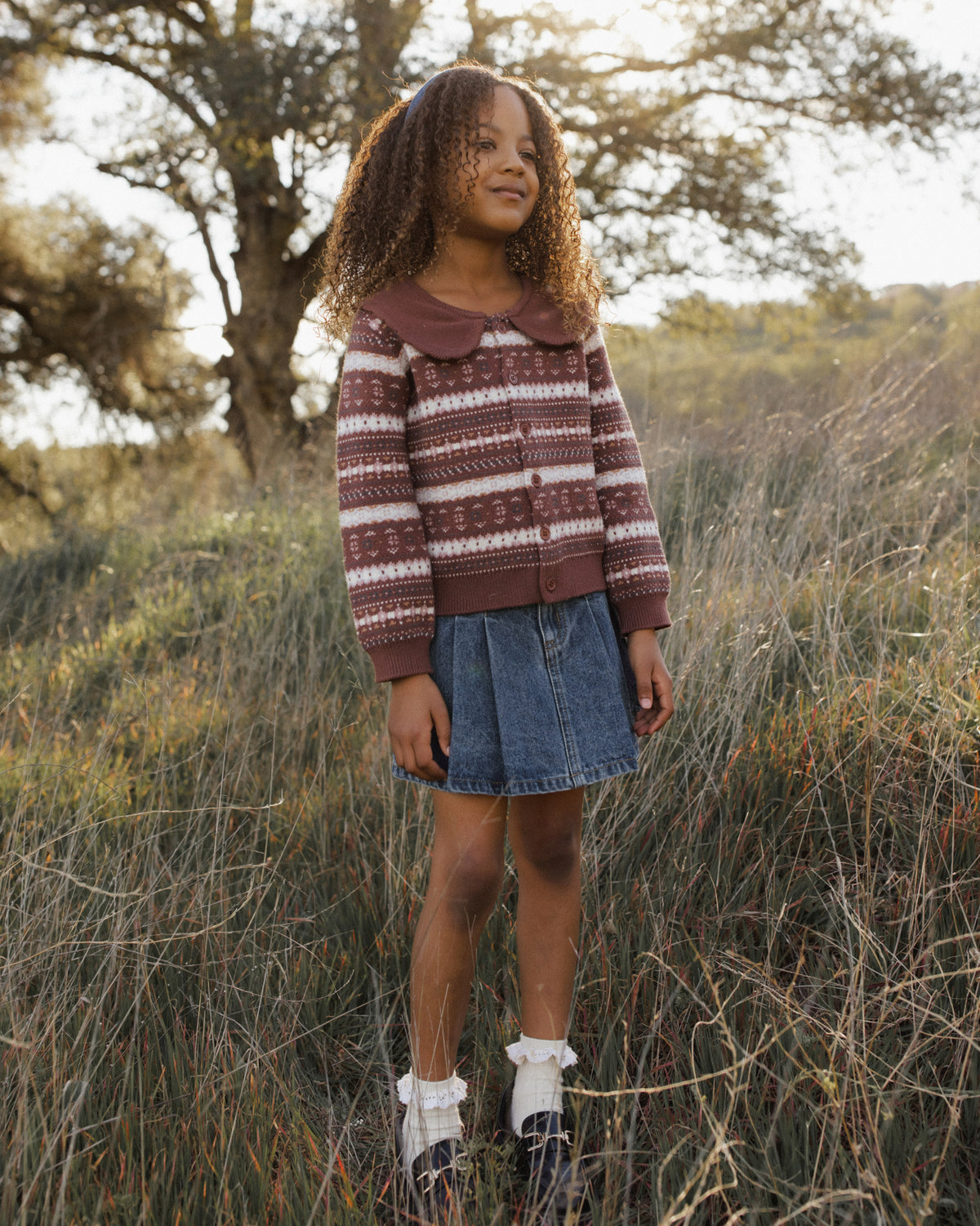 A young girl stands outdoors in a grassy field wearing a brown and white striped knit sweater, a denim skirt, white socks, and dark boots.