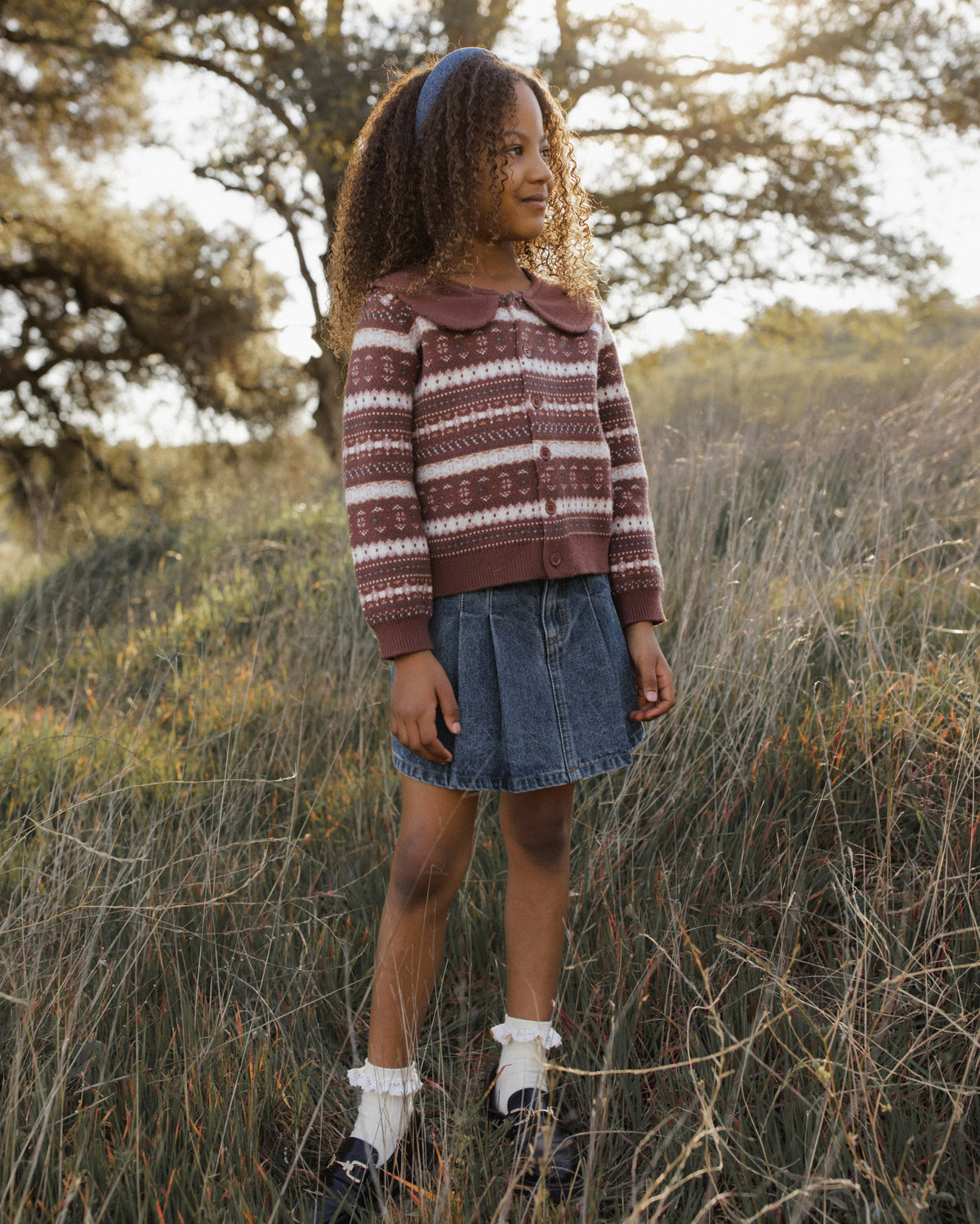 A young girl standing outdoors in a field of dry grass, wearing a burgundy and beige striped knit sweater, a denim mini skirt, white socks, and dark shoes.