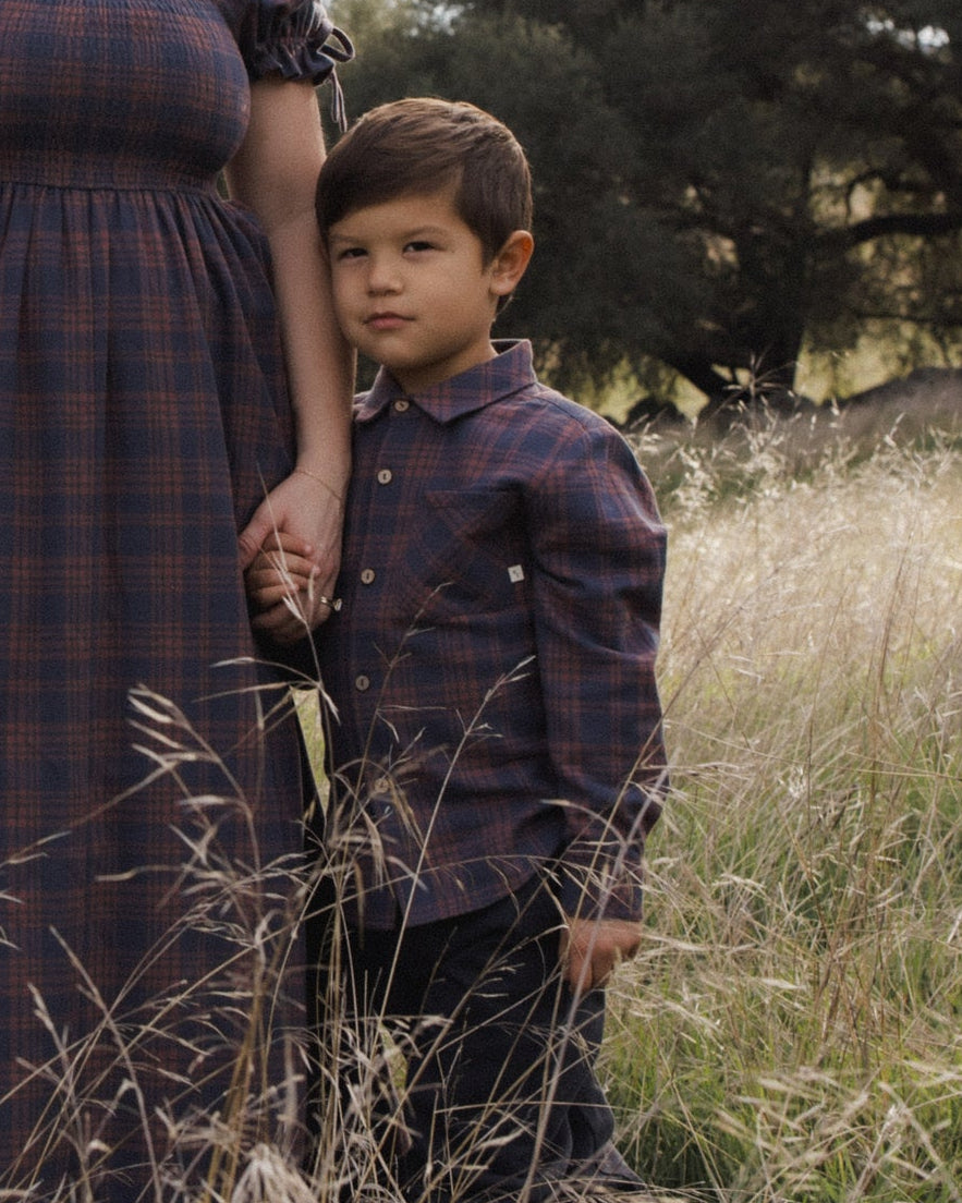 A young boy wearing a dark blue and brown plaid button-down shirt and dark pants stands in a grassy outdoor field next to an adult woman in a matching plaid dress.