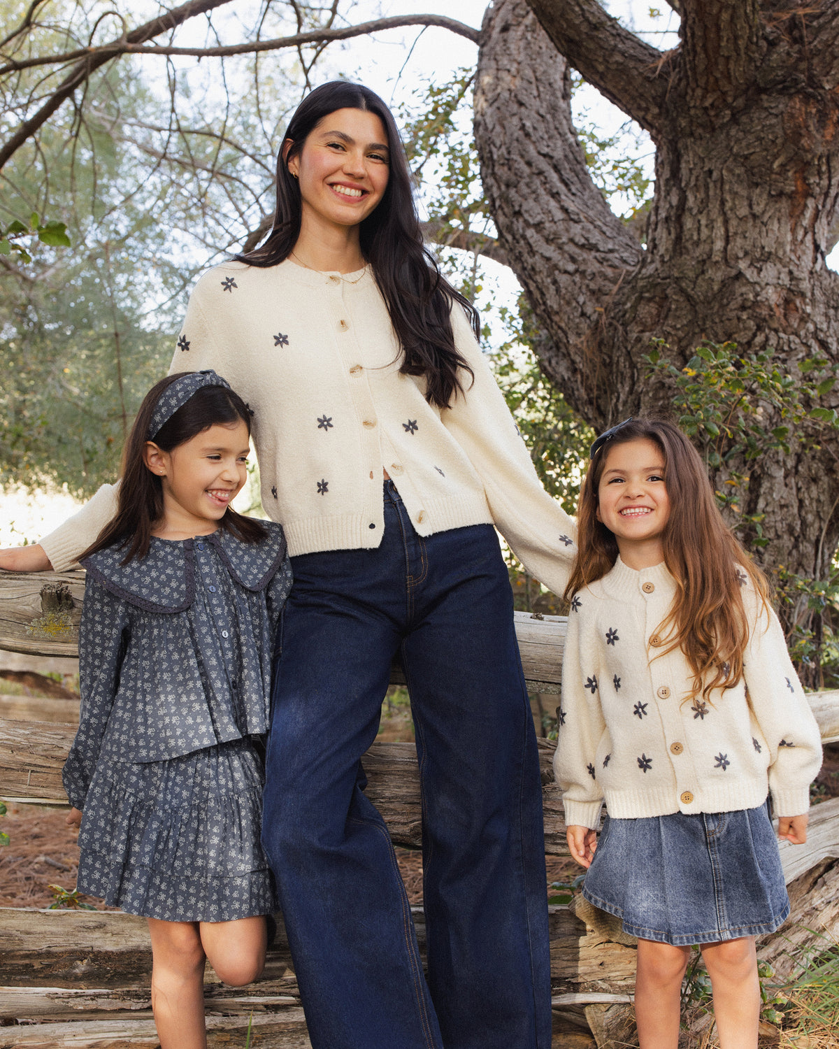 A woman and two young girls standing outdoors in front of a large tree, wearing coordinated knit clothing. The woman wears a cream knit top with small dark motifs and dark wide-leg pants, while the girls wear matching cream and gray outfits.