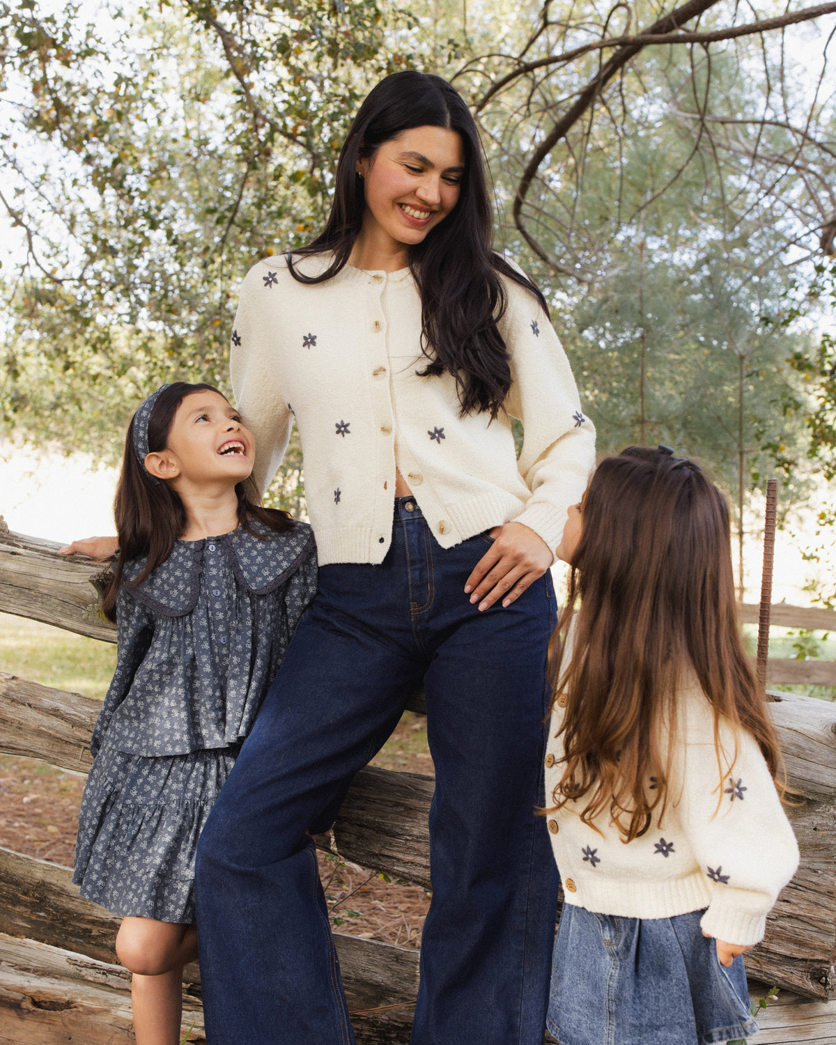 A woman and two young girls standing outdoors near a wooden fence and trees. The woman wears a cream cardigan with black polka dots and dark wide-leg pants, while the girls wear patterned dresses and tops.