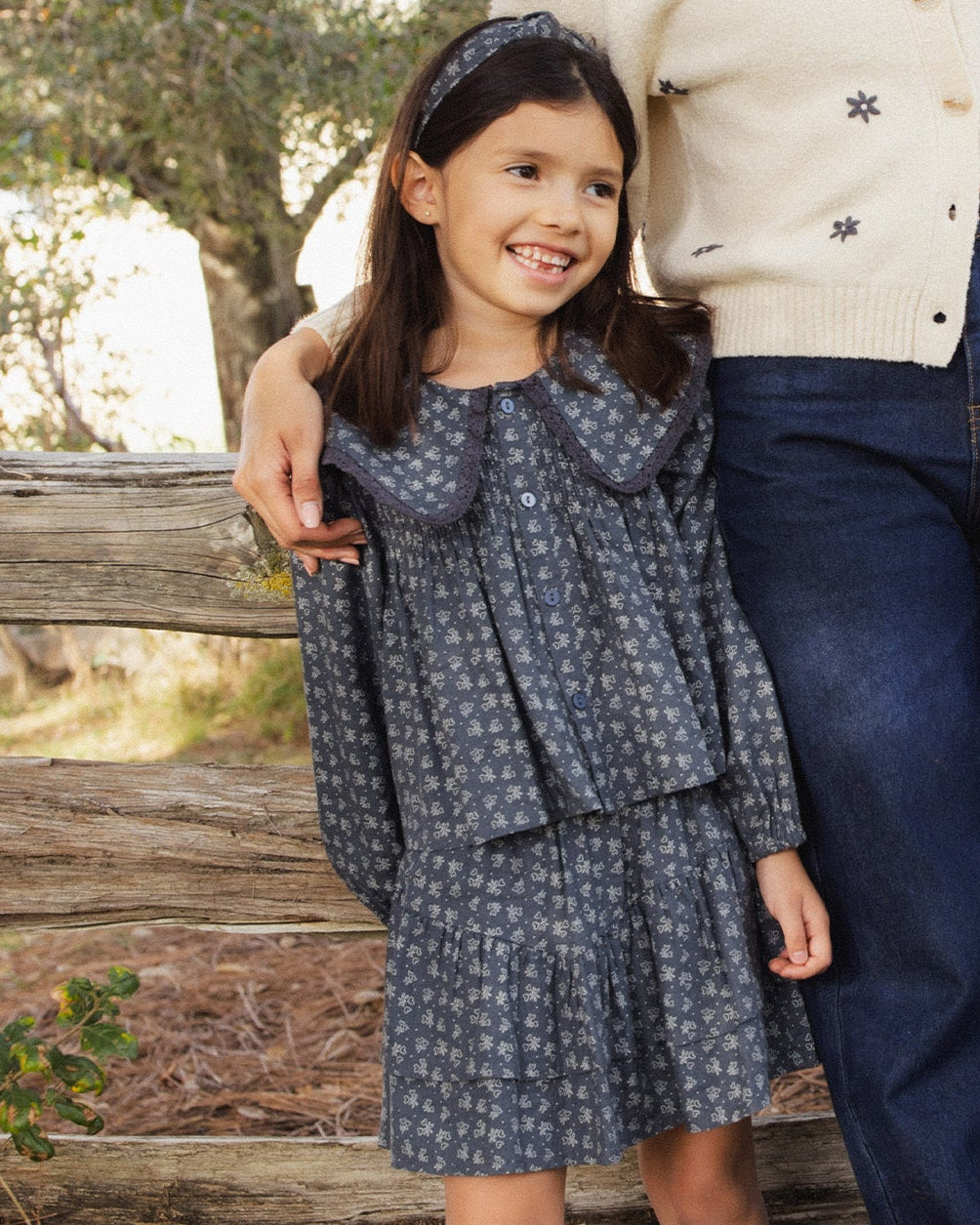 A young girl wearing a dark blue-gray long-sleeve dress with a small white floral pattern, a large Peter Pan collar, and a matching headband, standing outdoors next to an adult wearing a cream sweater and blue jeans.