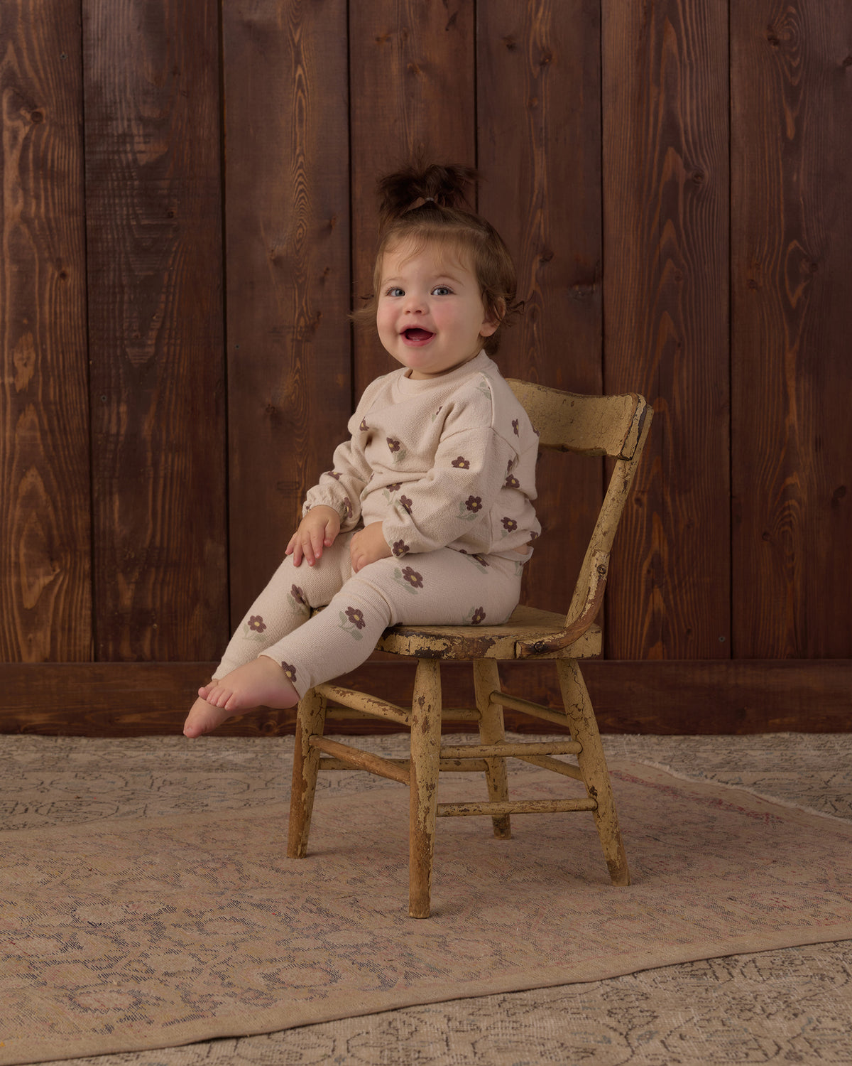 A toddler girl with a small hair bun sits on a rustic wooden chair, wearing a cream-colored long-sleeve pajama set with a brown floral pattern. The background consists of dark brown wooden wall panels and a patterned rug.