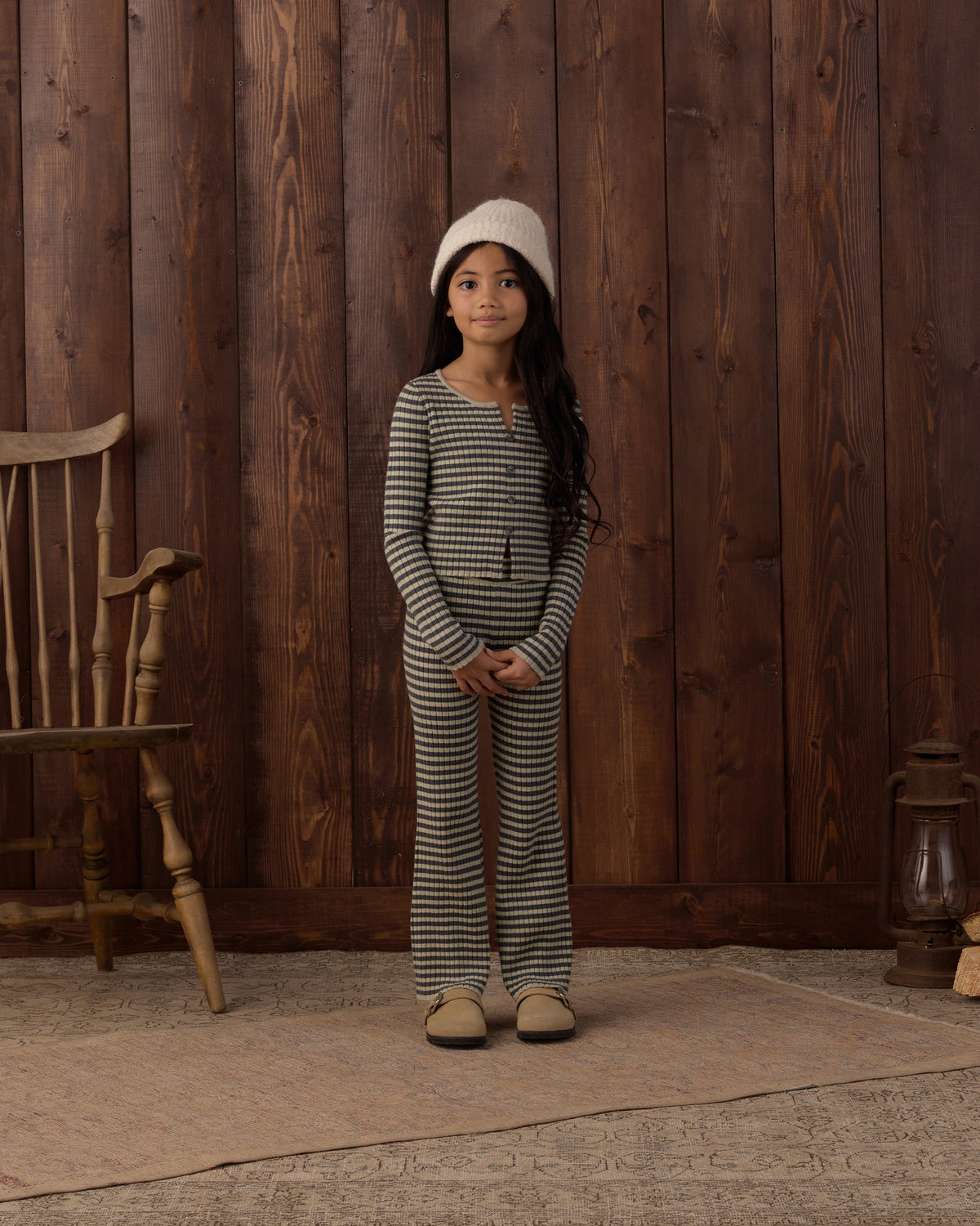 A young girl stands in front of a rustic dark wood plank wall wearing a matching two-piece striped lounge set with a white knit beanie and beige slippers.