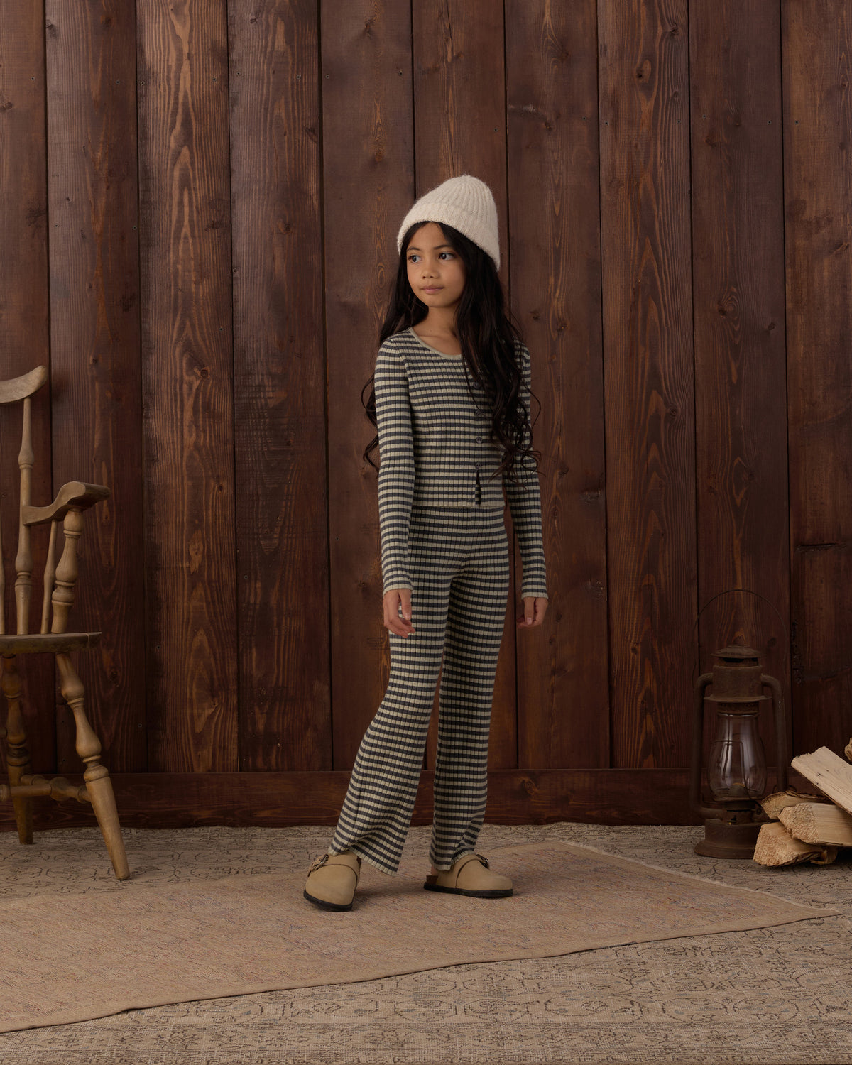 A young girl wearing a matching gray and white striped long-sleeve top and striped pants, styled with a cream knit beanie and beige shoes, standing in front of a rustic dark wood plank wall.