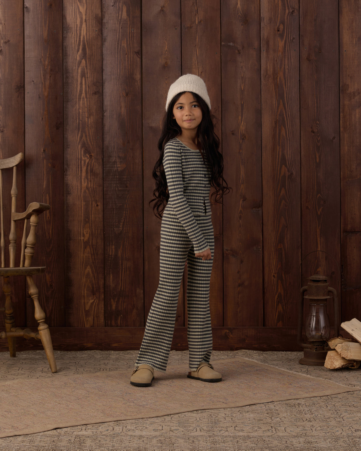 A young girl stands in front of a rustic dark wood plank wall wearing a matching long-sleeve striped one-piece outfit with a cream knit beanie and tan shoes.