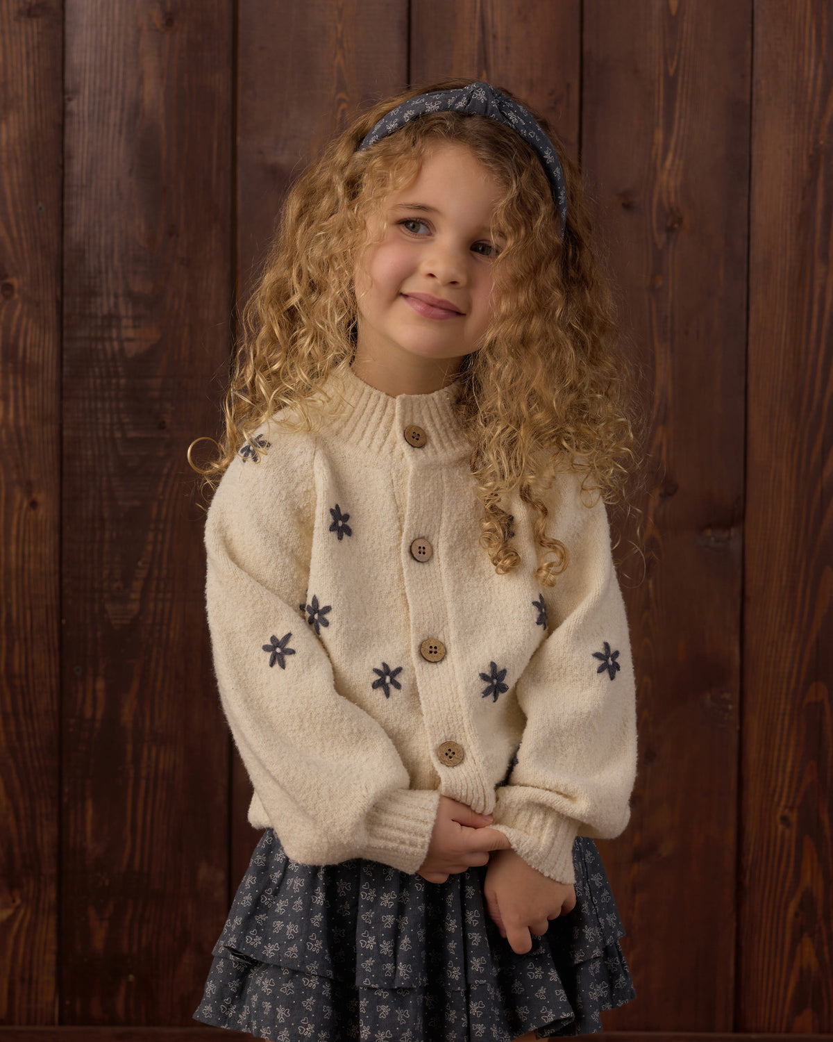 A young girl with curly blonde hair wearing a cream knit cardigan with dark blue embroidered snowflakes and a matching dark blue patterned skirt, standing against a dark wood plank background.