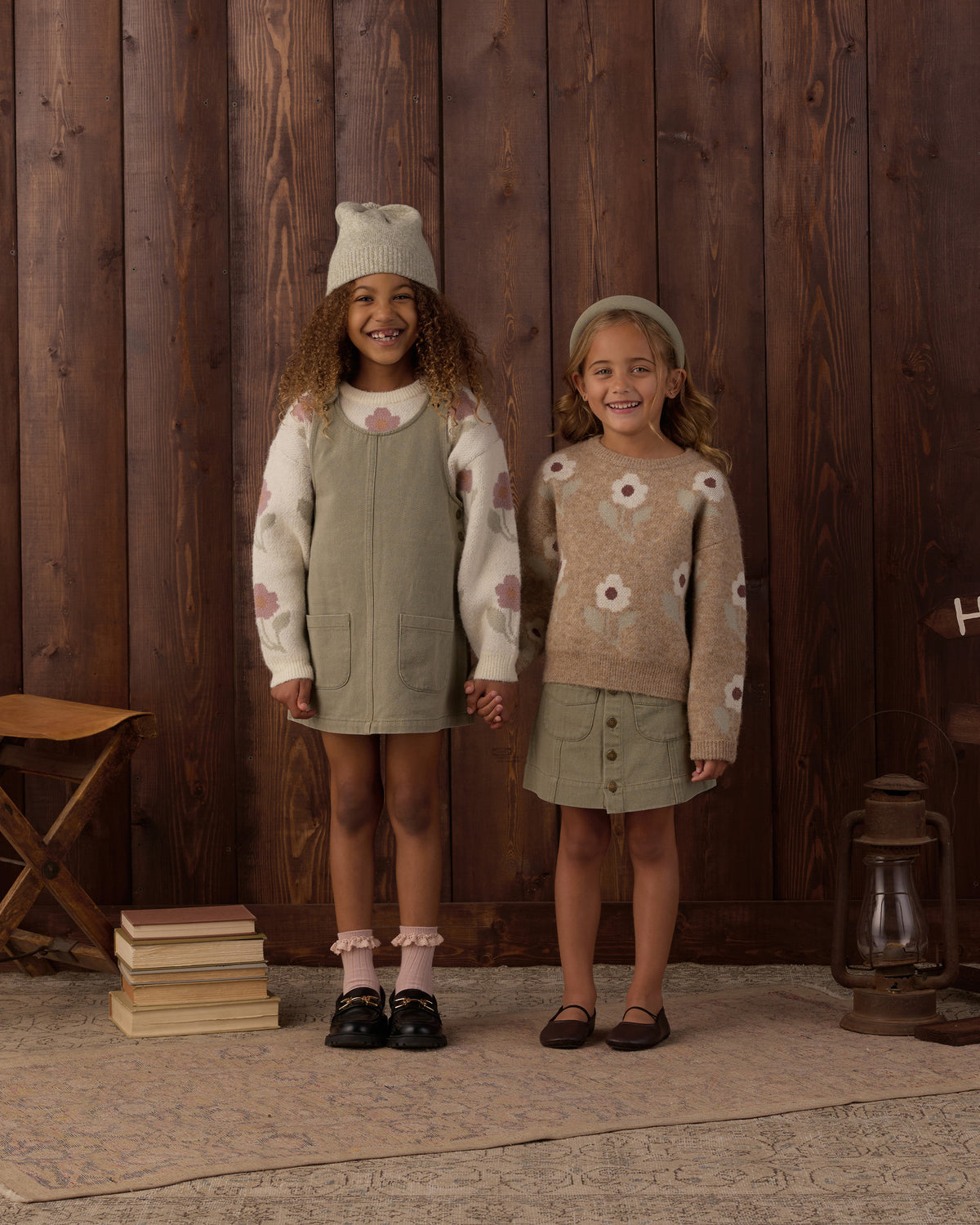 Two young girls modeling coordinated knit outfits in a rustic indoor setting with a dark wood plank wall. The girl on the left wears a cream knit sweater with pink floral motifs and a sage green pinafore dress, along with a matching cream knit hat.