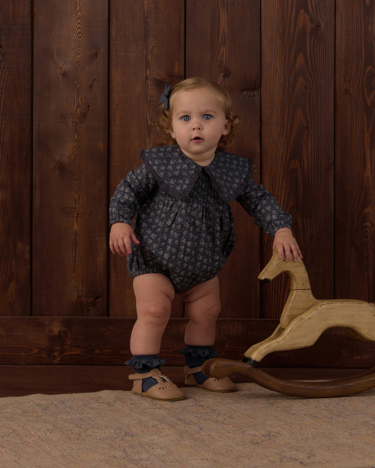A toddler stands indoors against a dark wood plank wall, wearing a dark navy long-sleeve romper with a small light pattern, ruffled collar, ruffled socks, and tan shoes. A wooden rocking horse is beside the child.