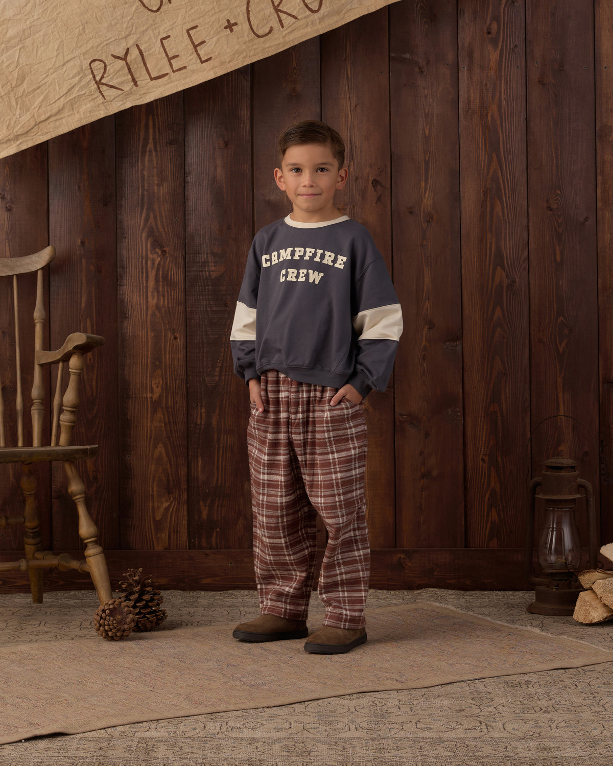 A young boy stands in front of a rustic dark wood plank wall wearing a navy blue long-sleeve sweatshirt with cream sleeves and 'CAMPFIRE CREW' printed in white on the chest, paired with brown and red plaid pajama pants.