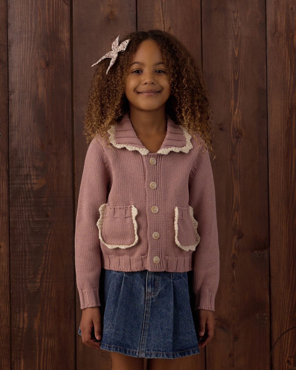 A young girl with curly hair wearing a dusty pink ribbed knit cardigan with cream ruffle trim and a denim skirt, standing against a dark wood plank background.
