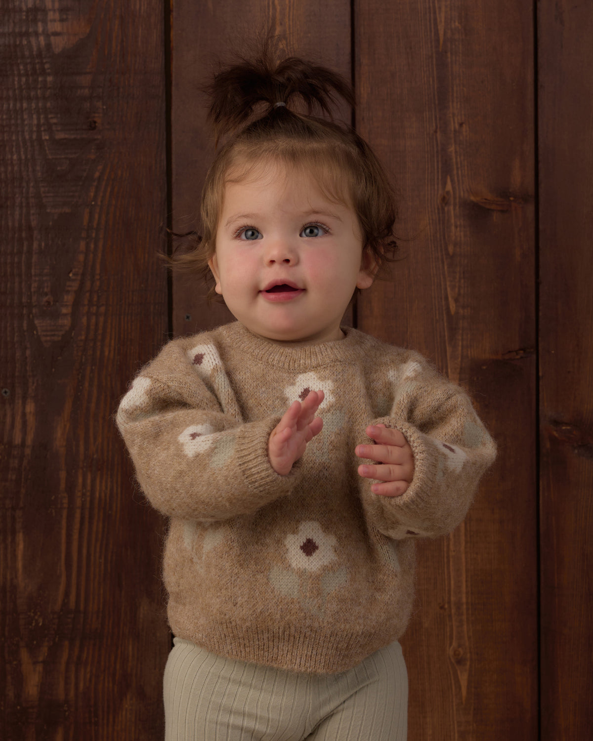 A toddler girl wearing a fuzzy beige knit sweater with white and brown floral patterns, standing in front of a dark wood plank background.