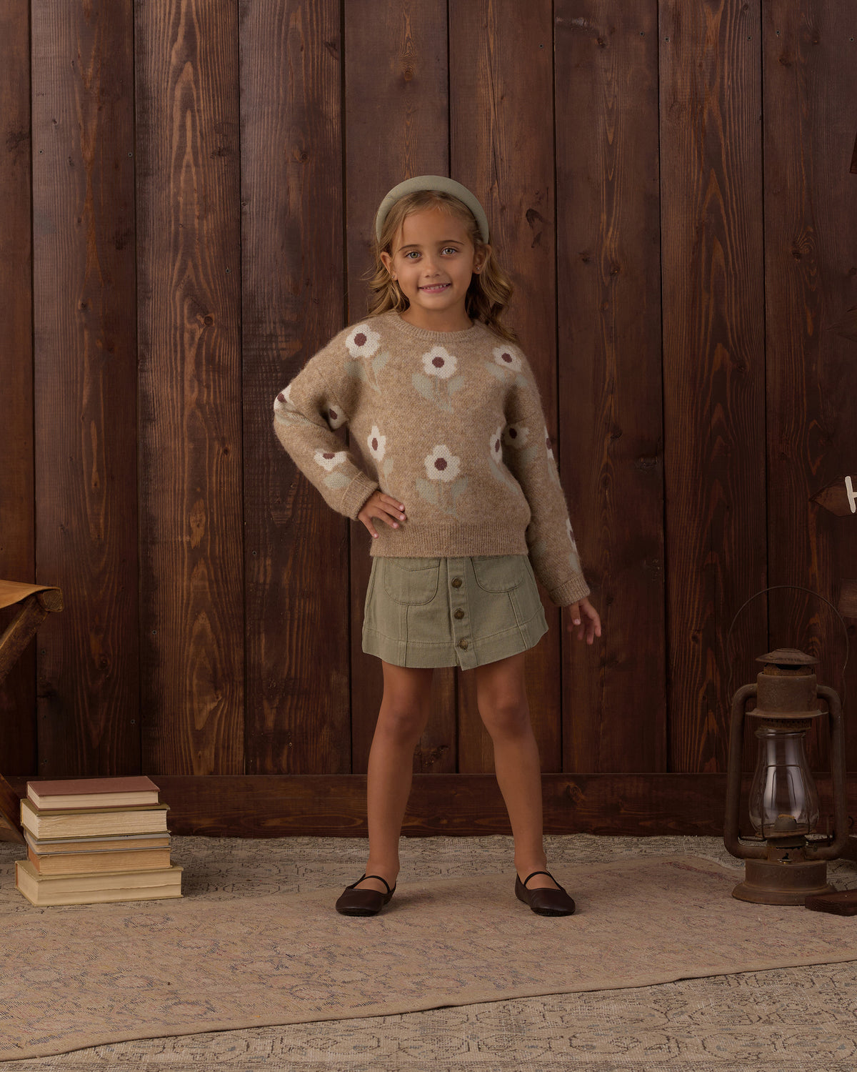 A young girl stands in front of a dark wood plank wall wearing a fuzzy beige sweater with white flower motifs, a sage green mini skirt, a matching green beanie, and black flats. A stack of books sits on the left, and a lantern rests on the right.