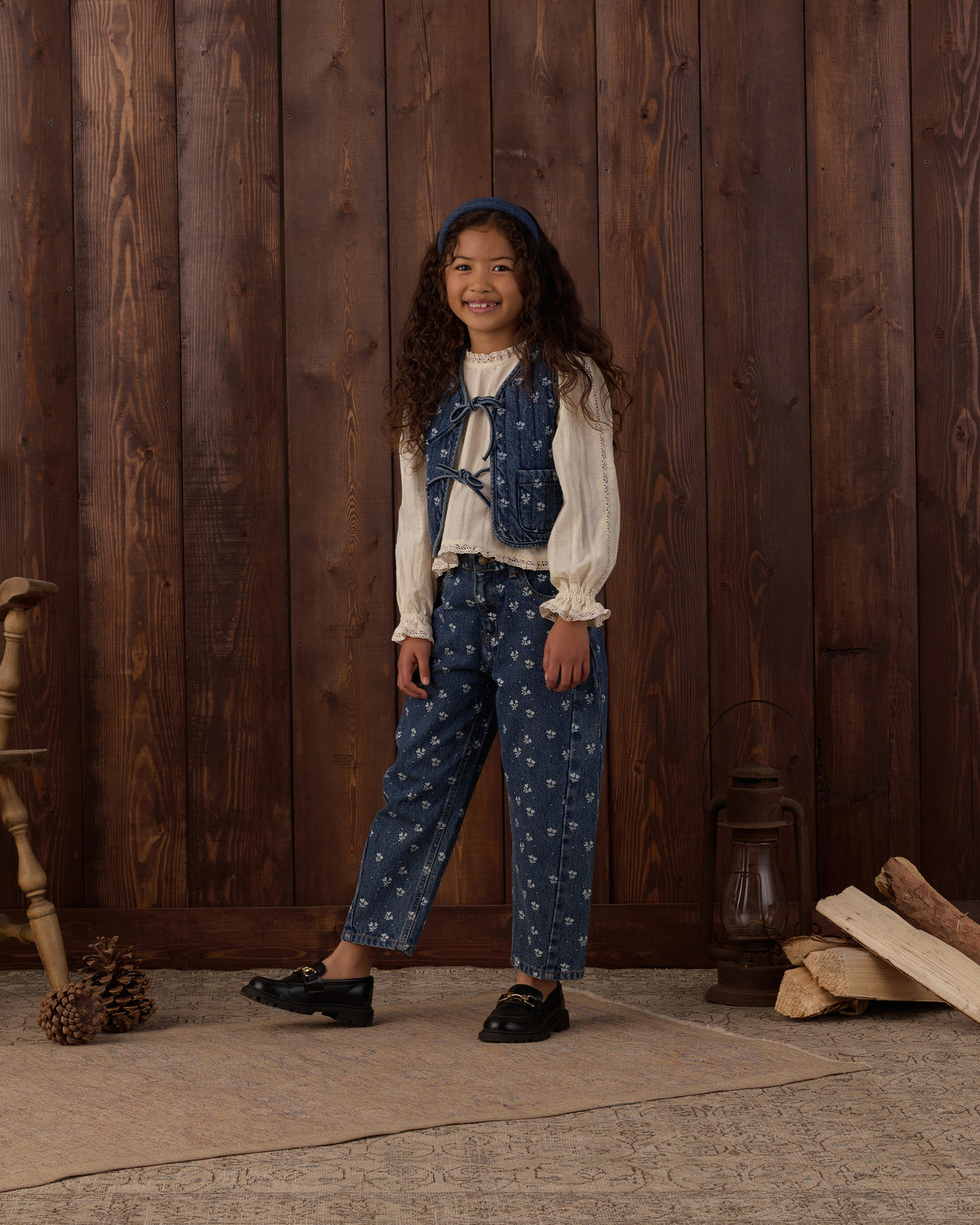 A young girl stands in front of a rustic dark wood plank wall wearing a vintage-style outfit consisting of a cream ruffled blouse, a blue floral vest, matching blue floral pants, and black shoes. The scene is styled with a wooden chair leg, pinecones, a lantern, and stacked logs on the floor.