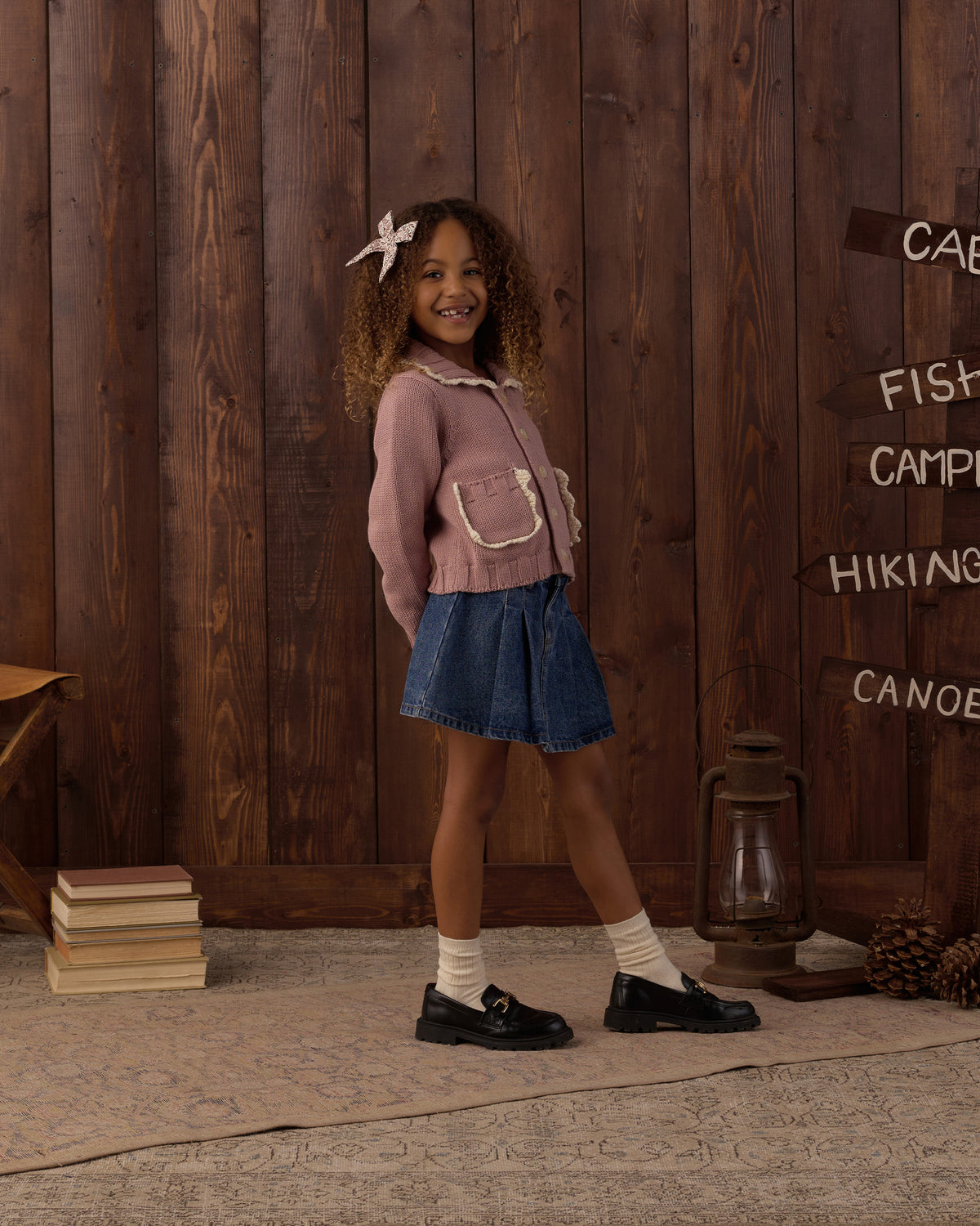 A young girl stands in front of a rustic dark wood plank wall wearing a dusty pink knit cardigan with white trim, a blue denim skirt, white socks, and black loafers. The background features a vintage-style lantern, a stack of books, and directional signs pointing to outdoor activities.