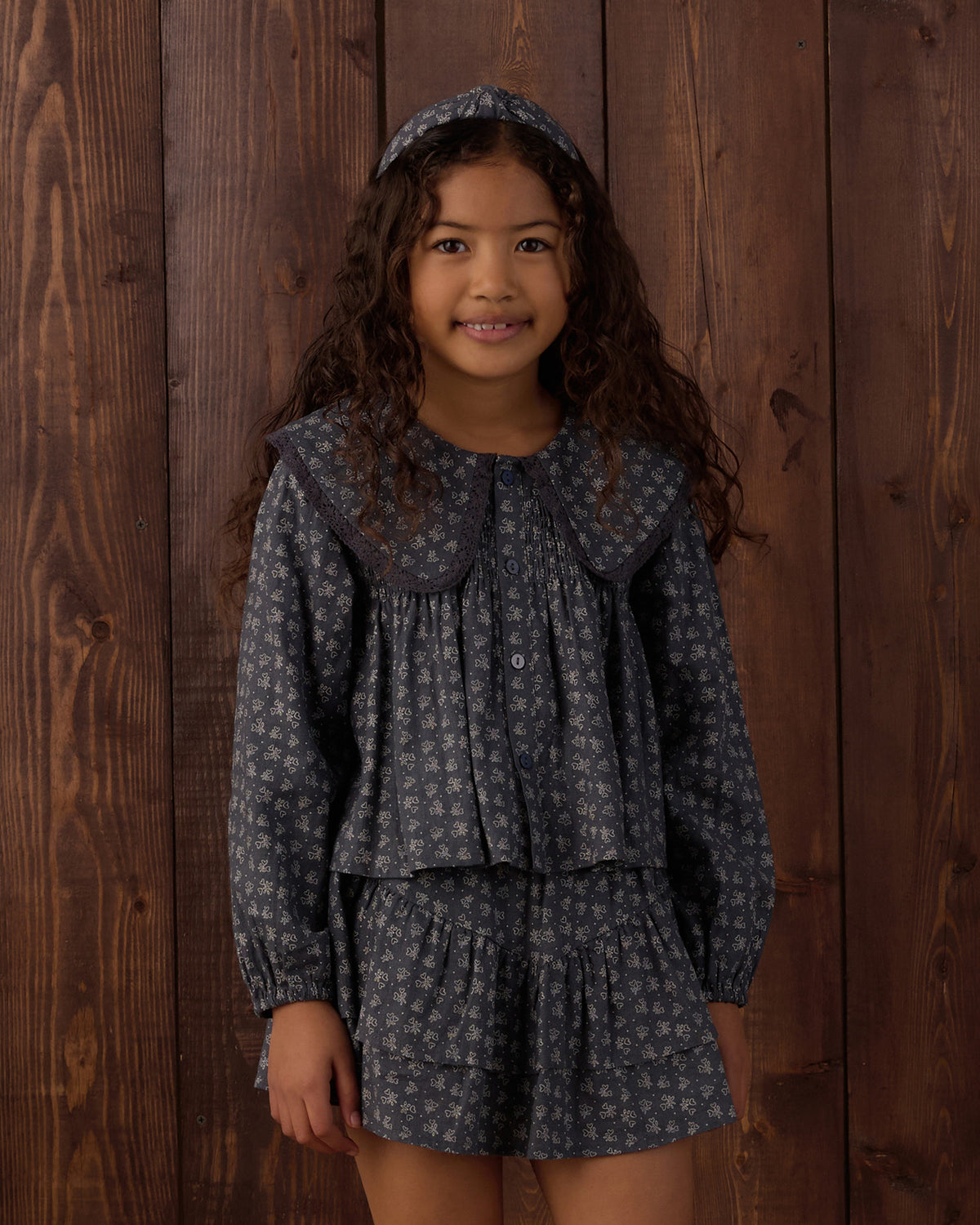 A young girl with long curly brown hair stands against a dark wood plank wall wearing a matching dark gray and white floral print outfit consisting of a long-sleeve button-front top with a large Peter Pan collar, a matching ruffled mini skirt, and a coordinating headband.