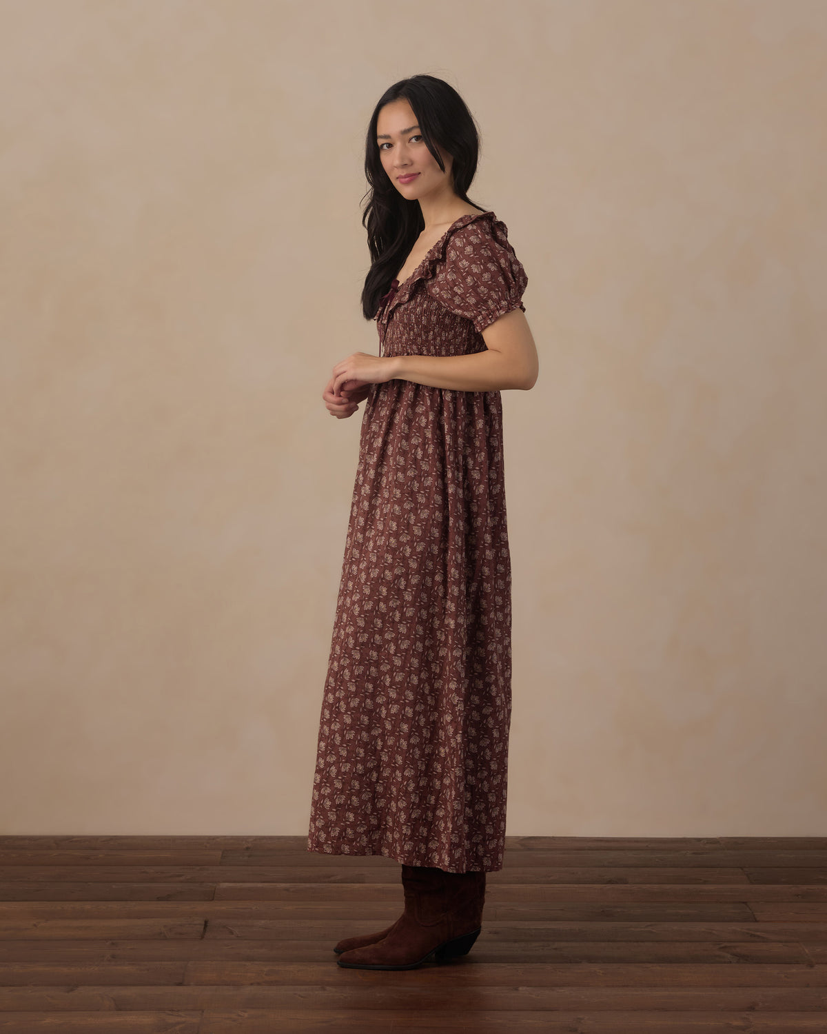 A woman modeling a brown short-sleeve maxi dress with a small floral pattern, standing against a neutral beige wall and wooden floor.
