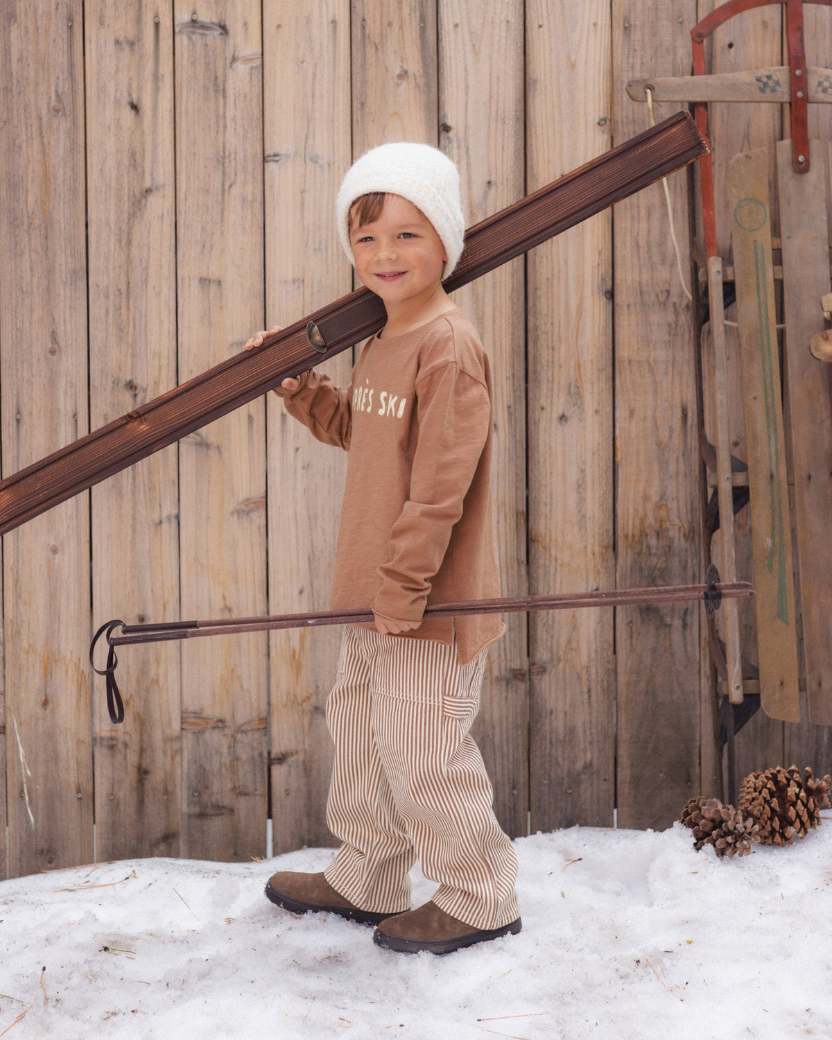 A young child wearing a brown long-sleeve shirt, beige pinstriped pants, and a white knit beanie stands in the snow holding a wooden sled against a rustic wooden fence.