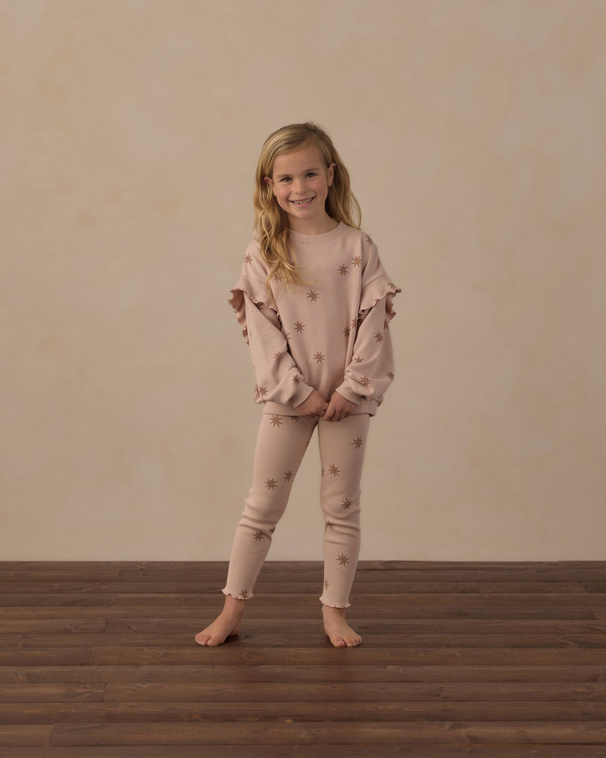 A young girl stands barefoot on a dark wood floor against a textured beige wall, wearing a matching dusty pink pajama set with star prints. The set includes a long-sleeve top with ruffled shoulders and matching leggings.