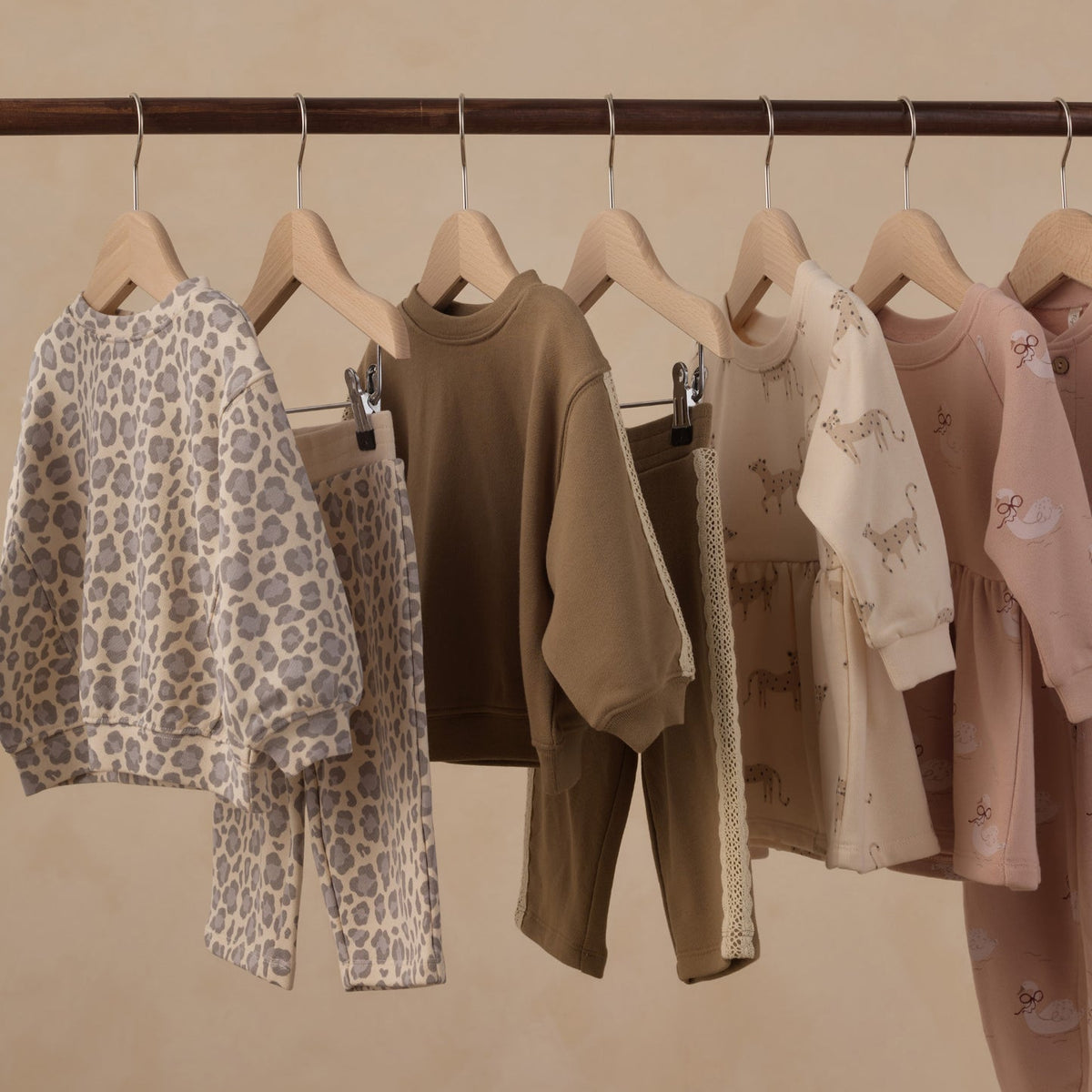 A set of children's two-piece pajama sets hanging on wooden hangers from a dark wooden rod against a warm beige wall. The set includes a beige and gray leopard print set, a solid brown set, a cream and beige animal print set, and a dusty pink set with small prints.