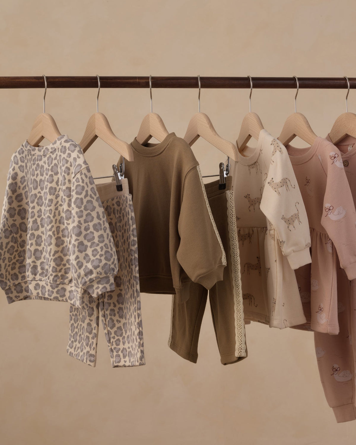 A set of children's two-piece pajama sets hanging on wooden hangers from a dark wooden rod against a warm beige wall. The set includes a beige and gray leopard print set, a solid brown set, a cream and beige animal print set, and a dusty pink set with small prints.