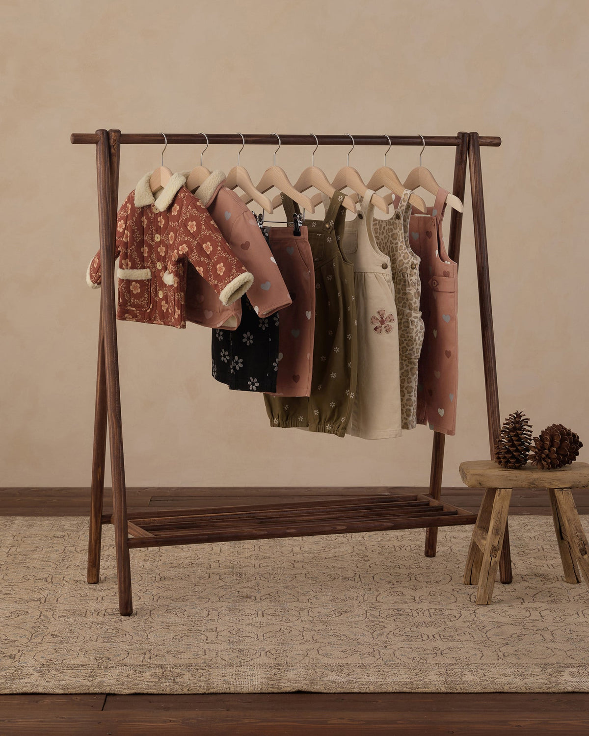 A clothing rack displaying multiple pairs of overall in different colors and patterns, including pink, olive green, beige, and black, hanging on wooden hangers against a warm beige background.