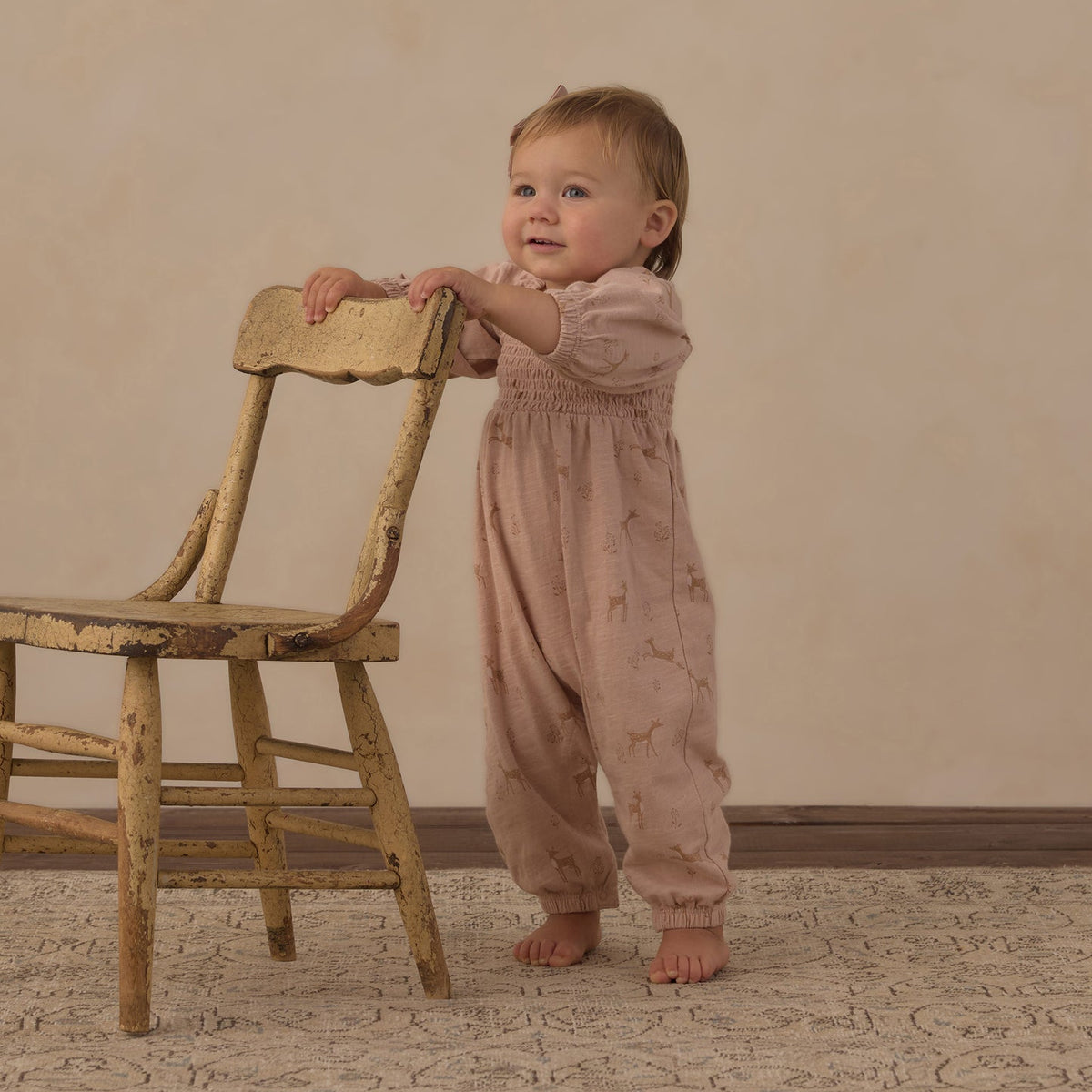 A baby standing on a patterned rug and leaning on a rustic wooden chair, wearing a dusty pink smocked jumpsuit with a subtle celestial print.