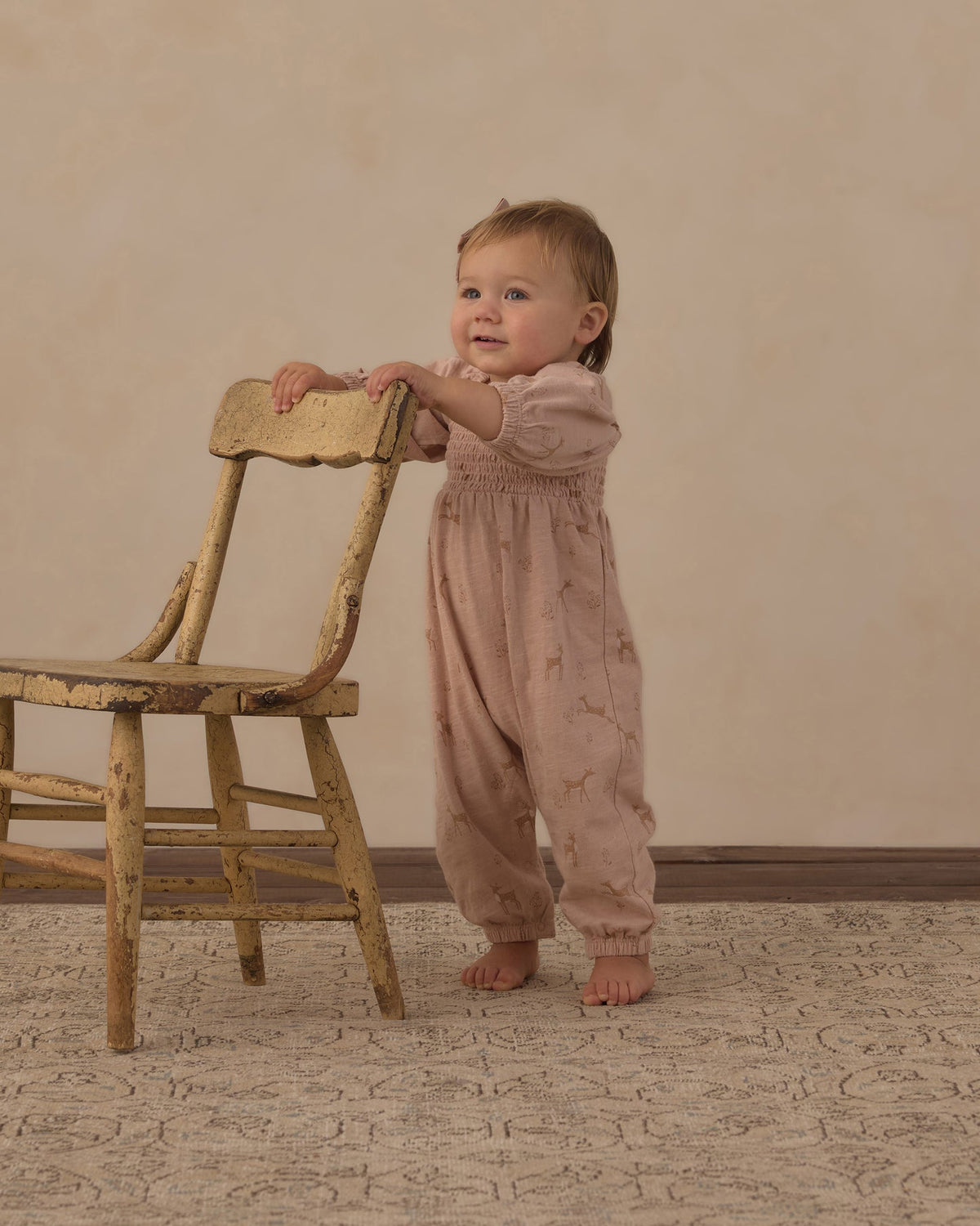 A baby standing on a patterned rug and leaning on a rustic wooden chair, wearing a dusty pink smocked jumpsuit with a subtle celestial print.