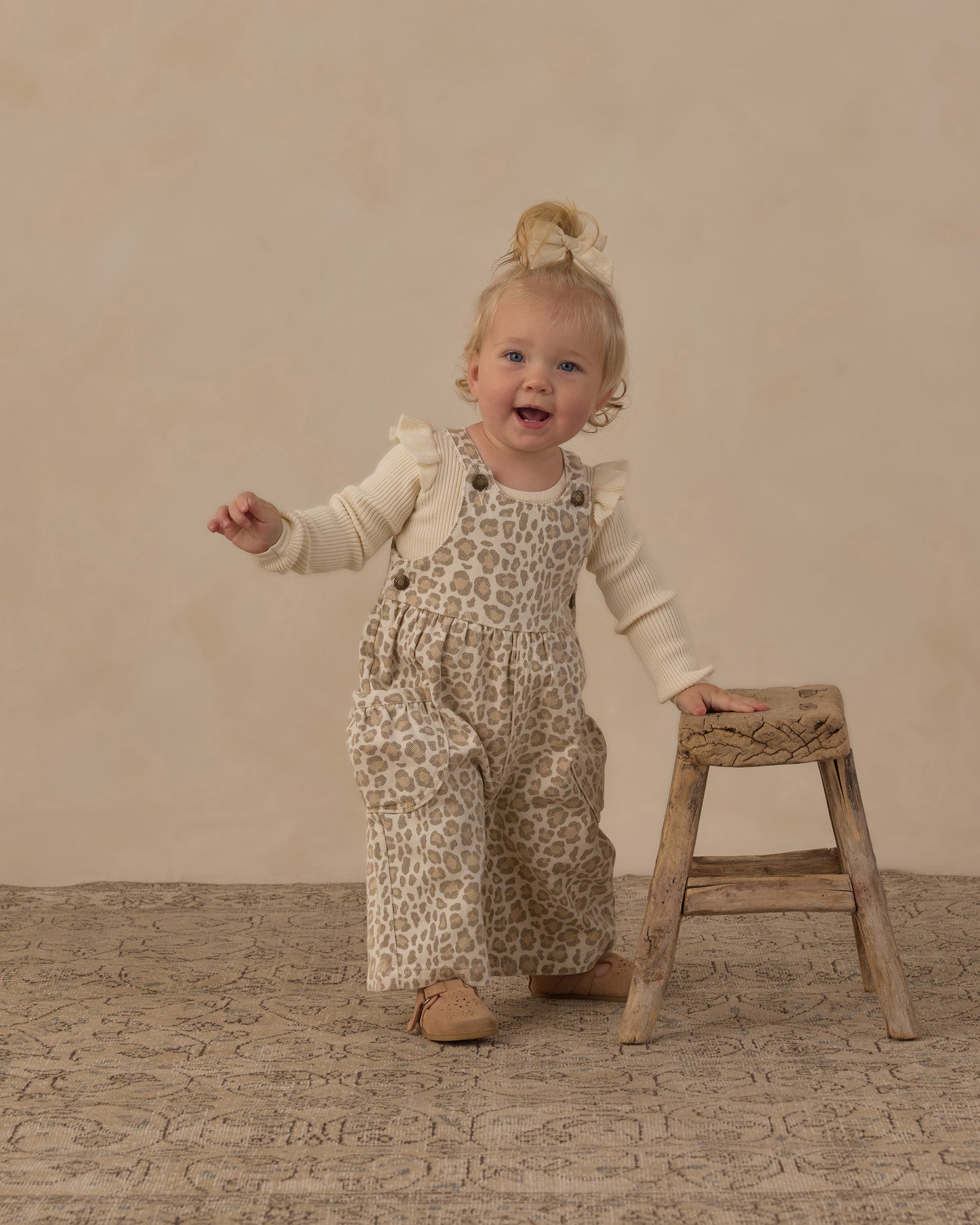 A toddler girl wearing beige and brown leopard-print overall with a cream long-sleeve shirt stands on a textured beige floor next to a small wooden stool, smiling and looking to the side.